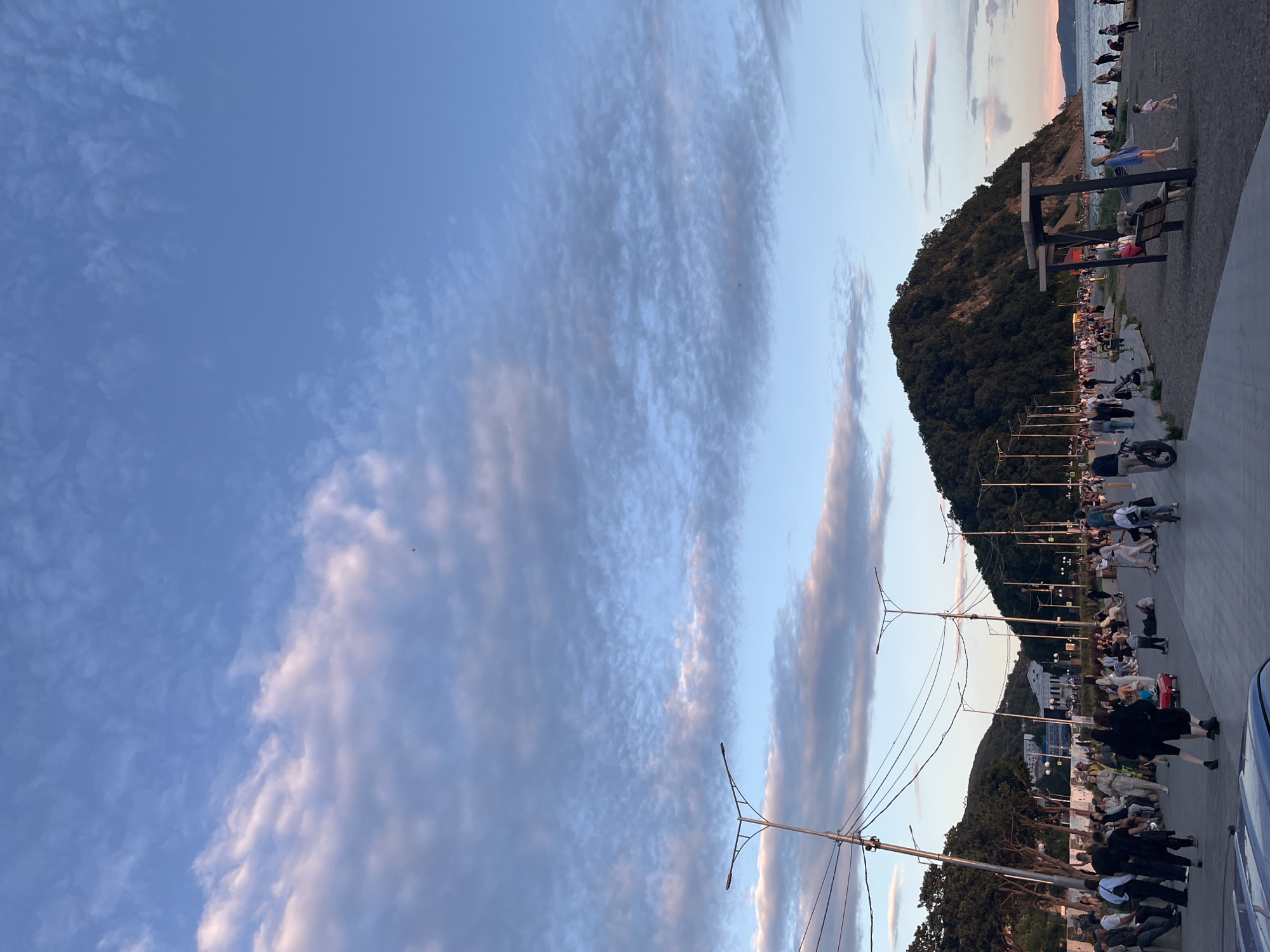 The image depicts a coastal promenade in Petropavlovsk-Kamchatsky, Russia, likely during the late afternoon or early evening, as indicated by the soft, warm lighting and the presence of a crowd enjoying outdoor activities. The scene is characterized by a wide, paved walkway bordering a beach and the sea. A prominent, densely forested hill dominates the right side of the frame, sloping down towards the water.
Numerous people are scattered across the promenade and the beach, engaged in various activities. Some are walking, others are seated on benches or the ground, and a few are riding bicycles or pushing strollers. There are also structures that appear to be part of the promenade's amenities, such as lampposts and a shaded seating area with benches. The sky above is a clear blue with scattered, wispy clouds, tinged with the pink and orange hues of sunset. The overall atmosphere suggests a lively and relaxed public space.