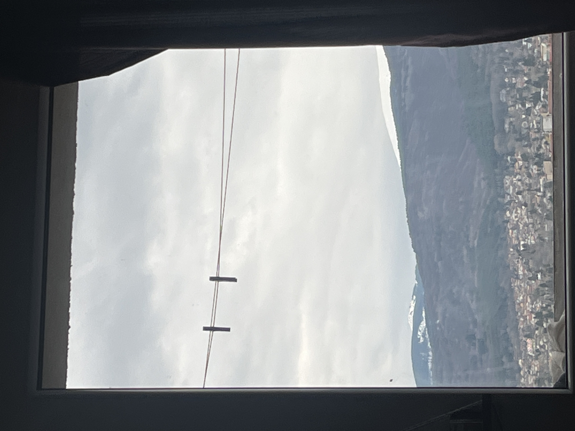 The image shows a view from a window, likely of a building in Sofia, Bulgaria. Outside the window, the sky is overcast with grey clouds. To the right of the window, a mountain is visible, with a settlement of buildings appearing at the base of the mountain. There are two parallel wires running vertically across the window frame, with two small, dark rectangular objects attached to the wires. The window frame itself is dark and indistinct in the foreground.
