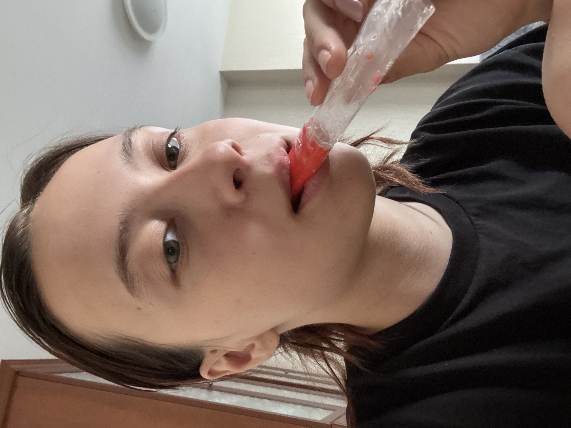 A woman is holding a red popsicle stick in her mouth. She is wearing a black t-shirt. The location is unknown.