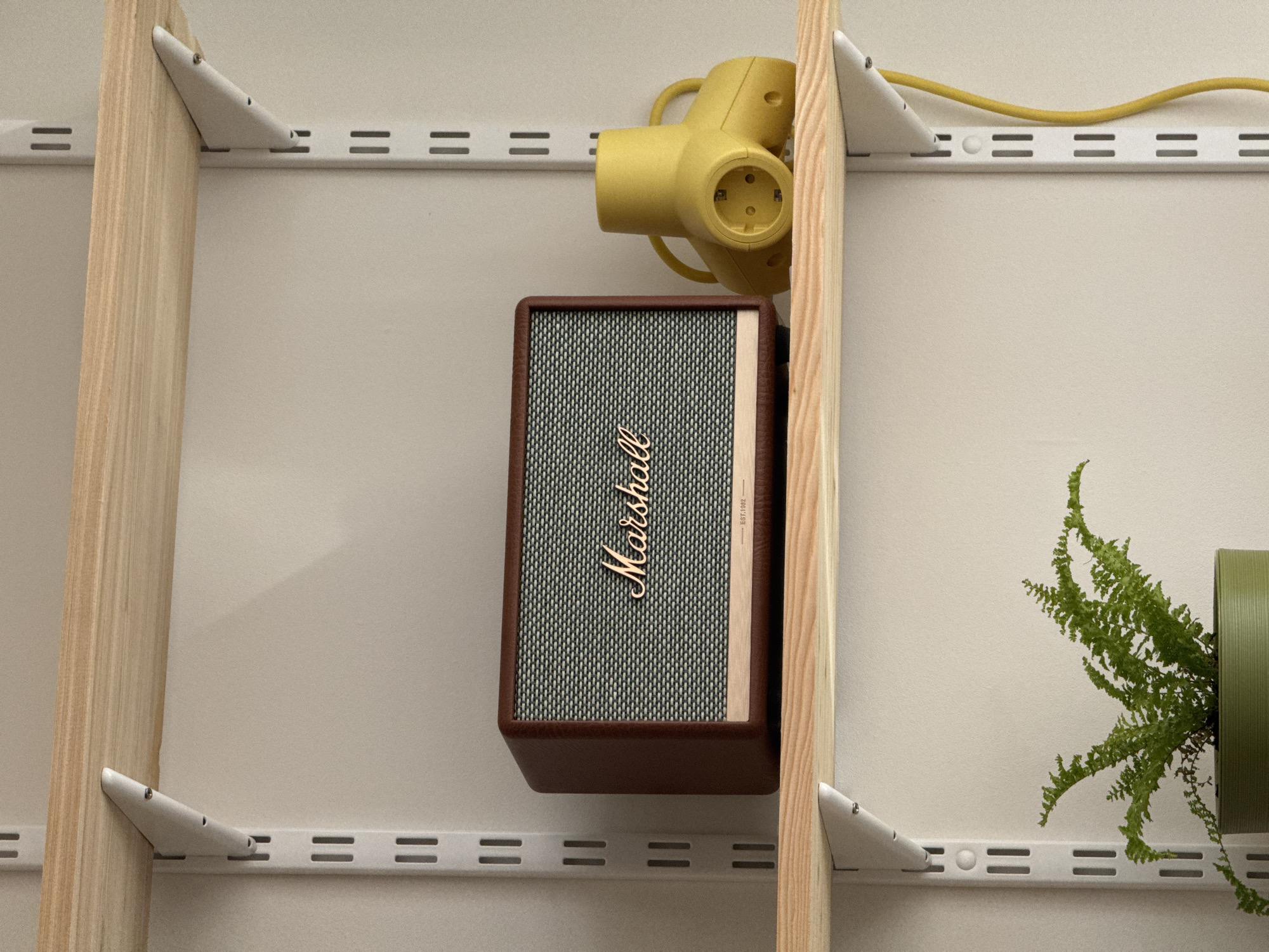 A close-up shot showcases a Marshall speaker nestled on a shelf, positioned beside a yellow power adapter. To the right, a green potted fern adds a touch of nature. The shelf itself is a functional structure made of light wood verticals with white brackets attaching it to a wall. The brackets and wall have evenly spaced parallel grooves for bracket adjustments.