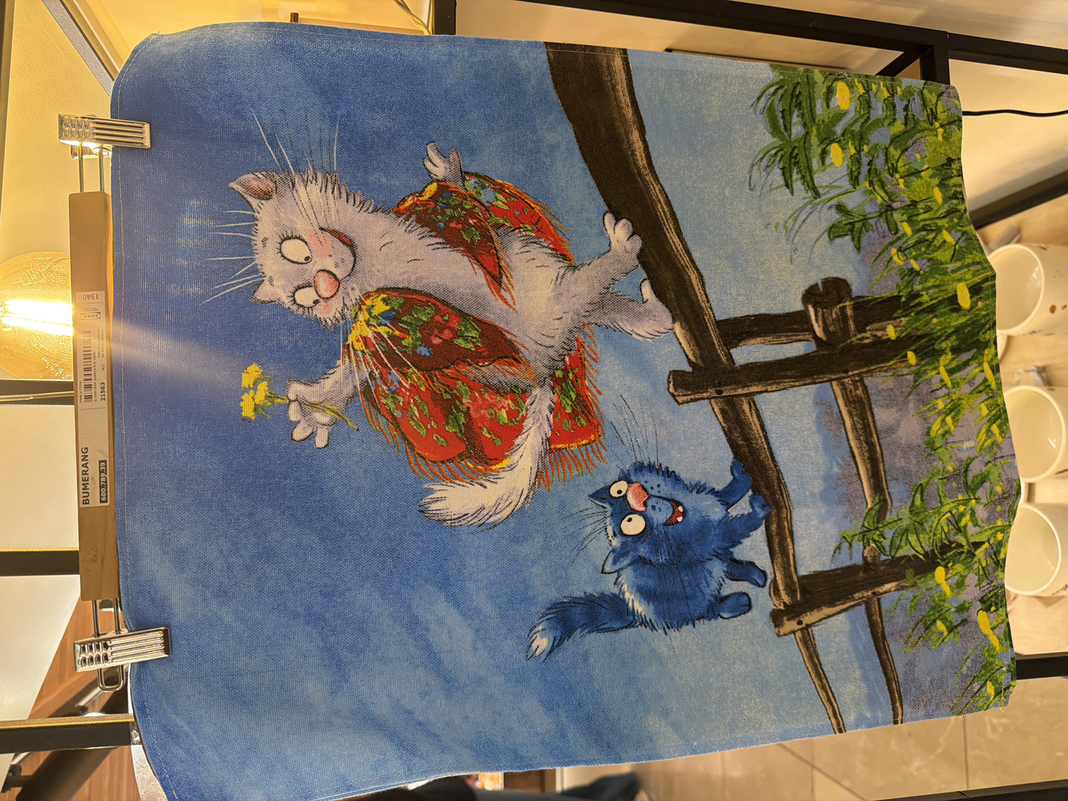 An image displays a fabric item, possibly a tea towel or small tapestry, featuring a whimsical cartoon illustration, held vertically by a wooden clothes hanger with metal clips. The photograph was taken in Saint Petersburg, Russia.
The illustration depicts two expressive cats against a bright blue sky. In the upper-left, a large, fluffy white and grey cat with wide, pink-rimmed eyes and rosy cheeks appears to be floating or flying. It holds a small bunch of yellow flowers in its left paw and wears a vibrant red garment adorned with green and yellow floral patterns, reminiscent of a traditional Russian folk art shawl or skirt. Its expression suggests wide-eyed surprise or delight.
Below and to the right, a smaller, fluffy blue cat with large, round eyes, white pupils, and an open mouth is shown clinging to a rustic wooden fence. Its body is partially obscured by the dark brown fence posts, and it looks upwards at the white cat with an expression of shock or alarm. Green foliage with small yellow elements, possibly flowers or fruit, is visible behind the fence on the right side of the illustration, contrasting with the blue sky.
A white label attached to the wooden hanger is visible on the left side of the image. Visible text on the label includes "IKEA", "BUMERANG", "21563", and "600.782.39". Additionally, "OPEL 1340" is discernable further up the label. The overall scene is a brightly colored, lighthearted cartoon depicting feline antics. The bright illumination suggests it is daytime. The texture of the fabric print is visible across the illustration.