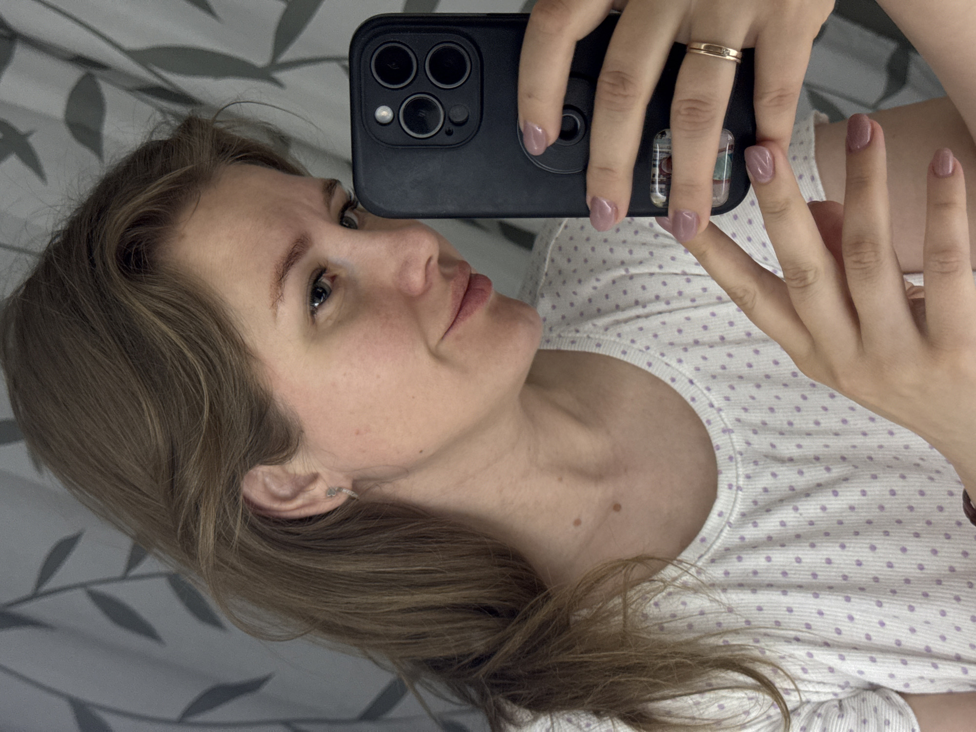 A young woman with light brown hair is taking a selfie with her black smartphone. She is wearing a white t-shirt with light purple polka dots and a gold ring on her left ring finger. The woman has light skin and her nails are painted with a light purple color. She is indoors, lying on a bed with a patterned sheet. The location is Gornyi (Dvurechenskoe S/P), Russia.