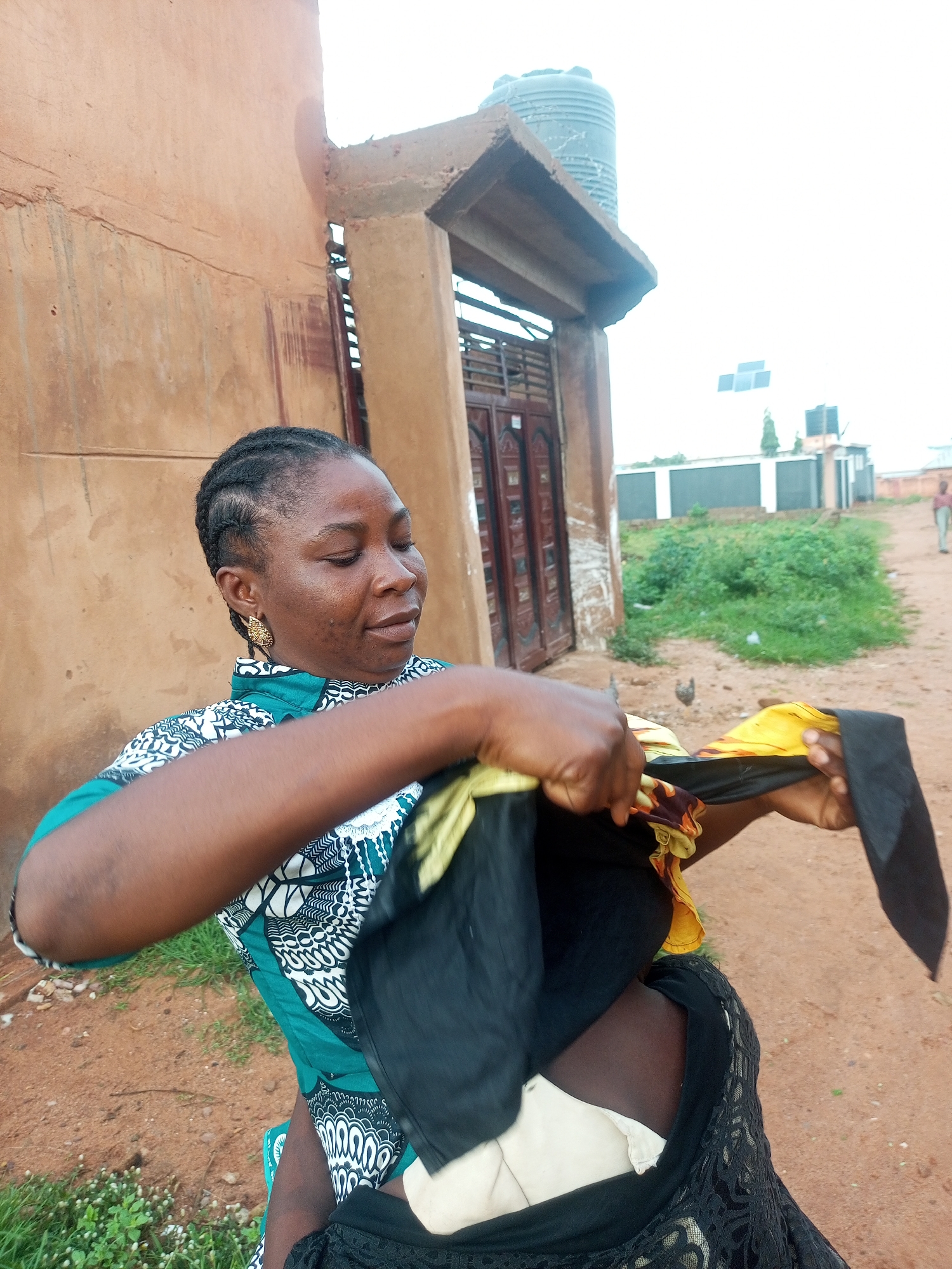 A medium shot captures a woman in Tempure, Nigeria, with her head and torso in the foreground. She has braided dark hair and is wearing a green, white, and black patterned top and large gold earrings. Her hands are busy folding a black garment with yellow and red floral print accents. Her eyes are closed, and her expression suggests concentration or relaxation. Behind her, a rustic, earth-toned building with an arched entryway is visible. To the right of the building, a large grey water tank sits atop a concrete structure, and in the distance, solar panels are mounted on a rooftop. The ground is a dusty reddish-brown, with patches of green grass and small white flowers. A narrow dirt path extends into the background, where other buildings and a person can be seen in the hazy distance under an overcast sky. A chicken is visible in the middle ground.