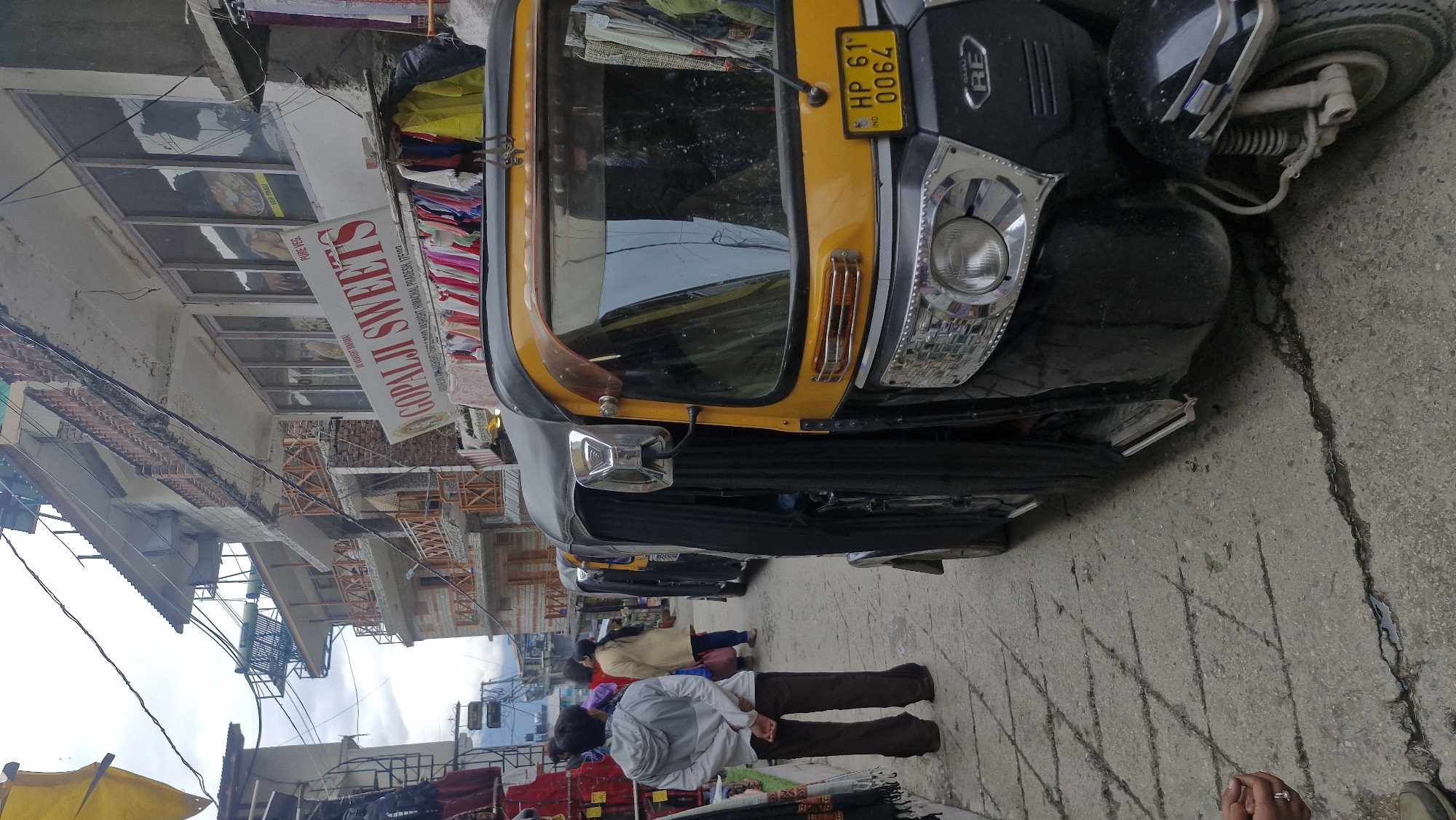 In Vashisht, India, a street scene unfolds with a prominent yellow and black auto-rickshaw parked on a cracked, grey asphalt road. The auto-rickshaw is angled towards the viewer, revealing its front details including a yellow license plate reading "HP 61 0064". To the left of the auto-rickshaw, a young person wearing a grey hooded jacket and brown pants stands with their back to the camera, hands behind their back. Further down the street, a woman in a beige coat and dark pants is visible. Above the auto-rickshaw, a sign reads "GOPALJI SWEETS". Buildings with balconies line the street, displaying various wares and advertisements. The sky above is overcast, suggesting a cloudy day.