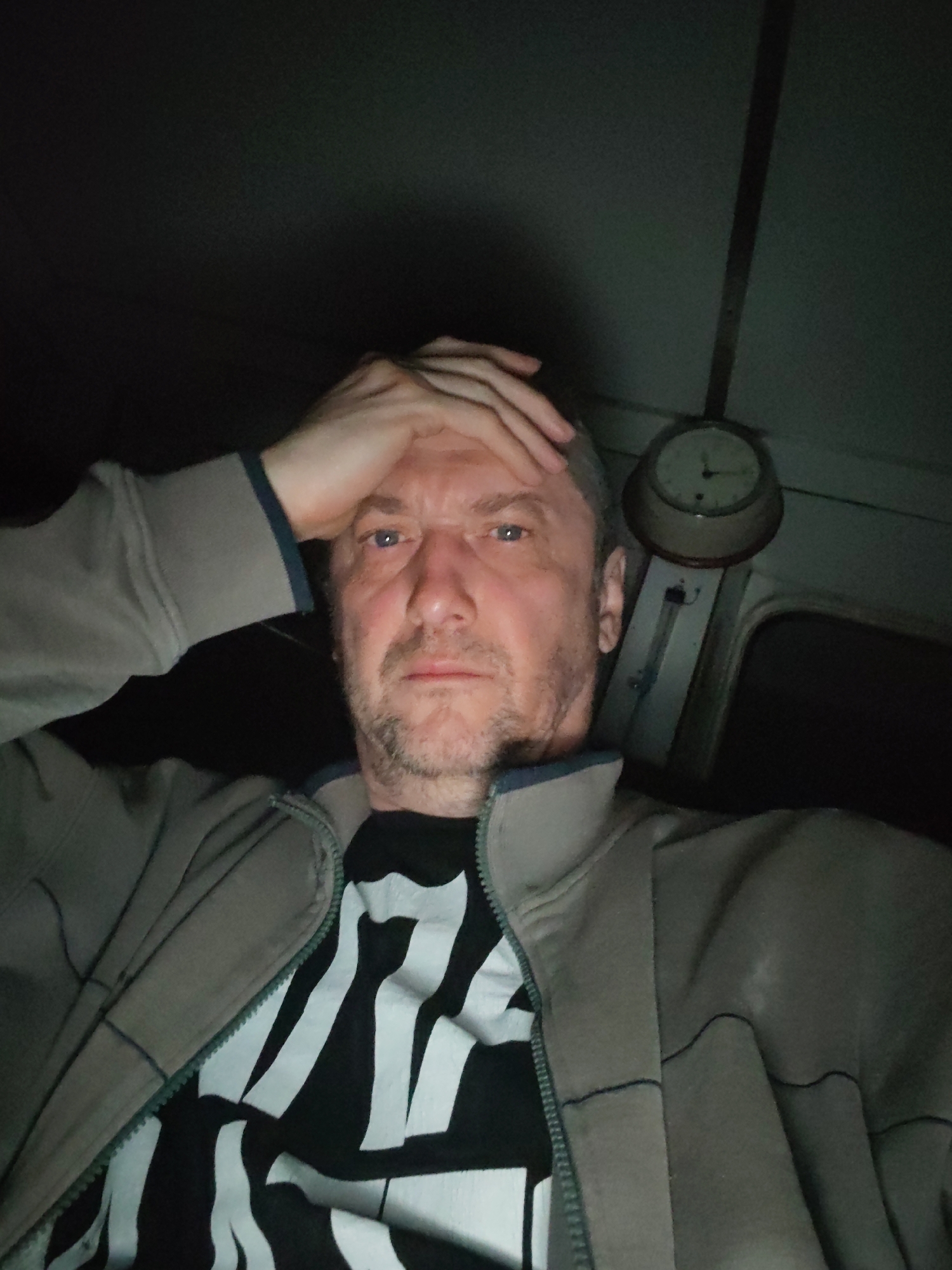 A middle-aged man with blue eyes, stubble, and fair skin is looking directly at the camera with a concerned expression. He has his right hand resting on his forehead. He is wearing a grey jacket with a dark collar over a black and white patterned t-shirt. Above and behind the man to the right is a round clock hanging on what appears to be a wall. The environment is dimly lit. There is no information available in the image to indicate its location in the city of Bol'shie Mkhi, Russia.