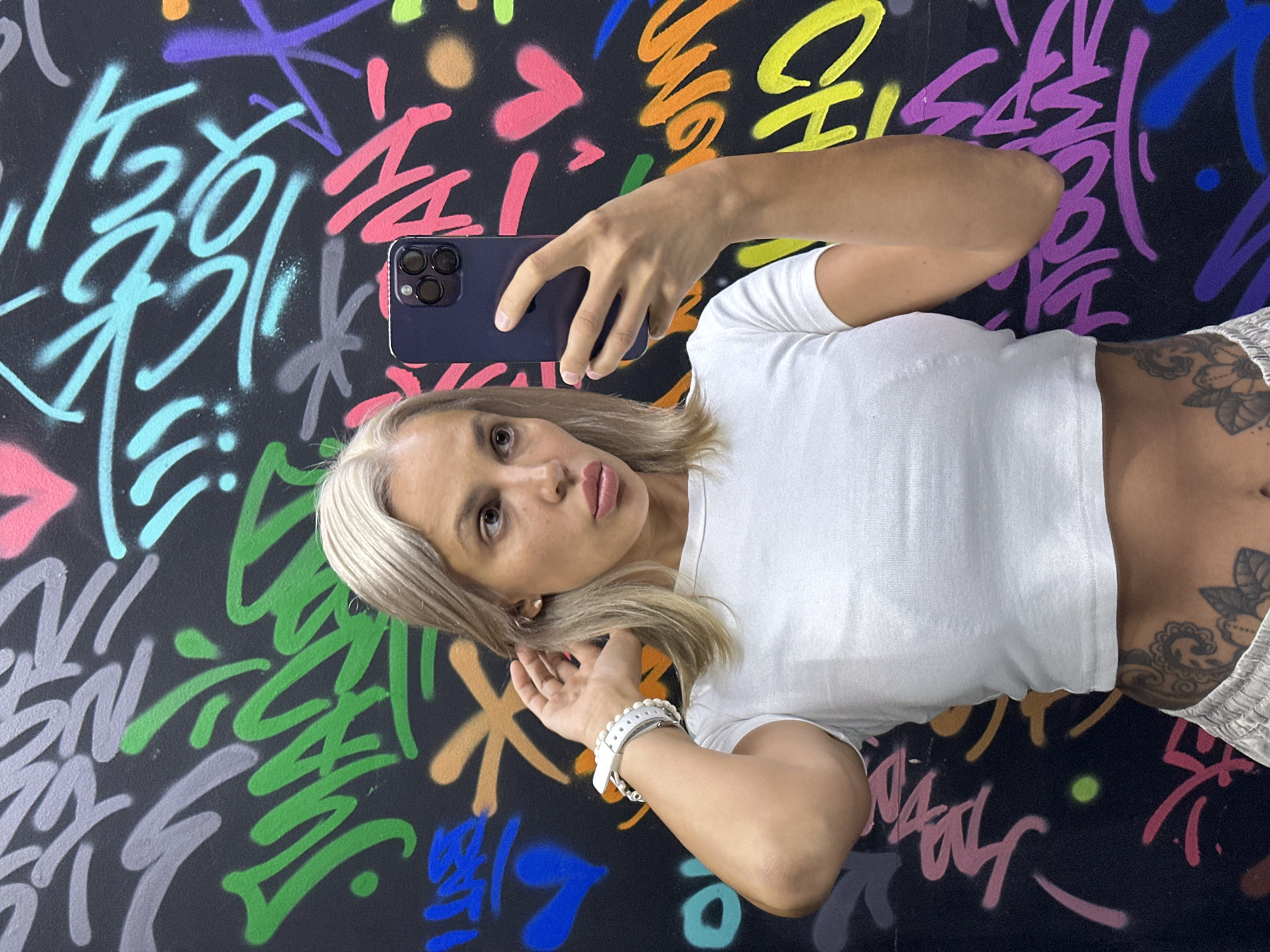 A young woman with blonde hair tied to her ear is taking a selfie against a black wall covered in colorful graffiti. She is wearing a white crop top and grey sweatpants, with a tattoo visible on her abdomen. She has a white watch on her left wrist. The graffiti on the wall features various colors and lettering, including pink, green, blue, yellow, and purple. The background suggests an urban setting, possibly a street art mural in Kuta, Indonesia.