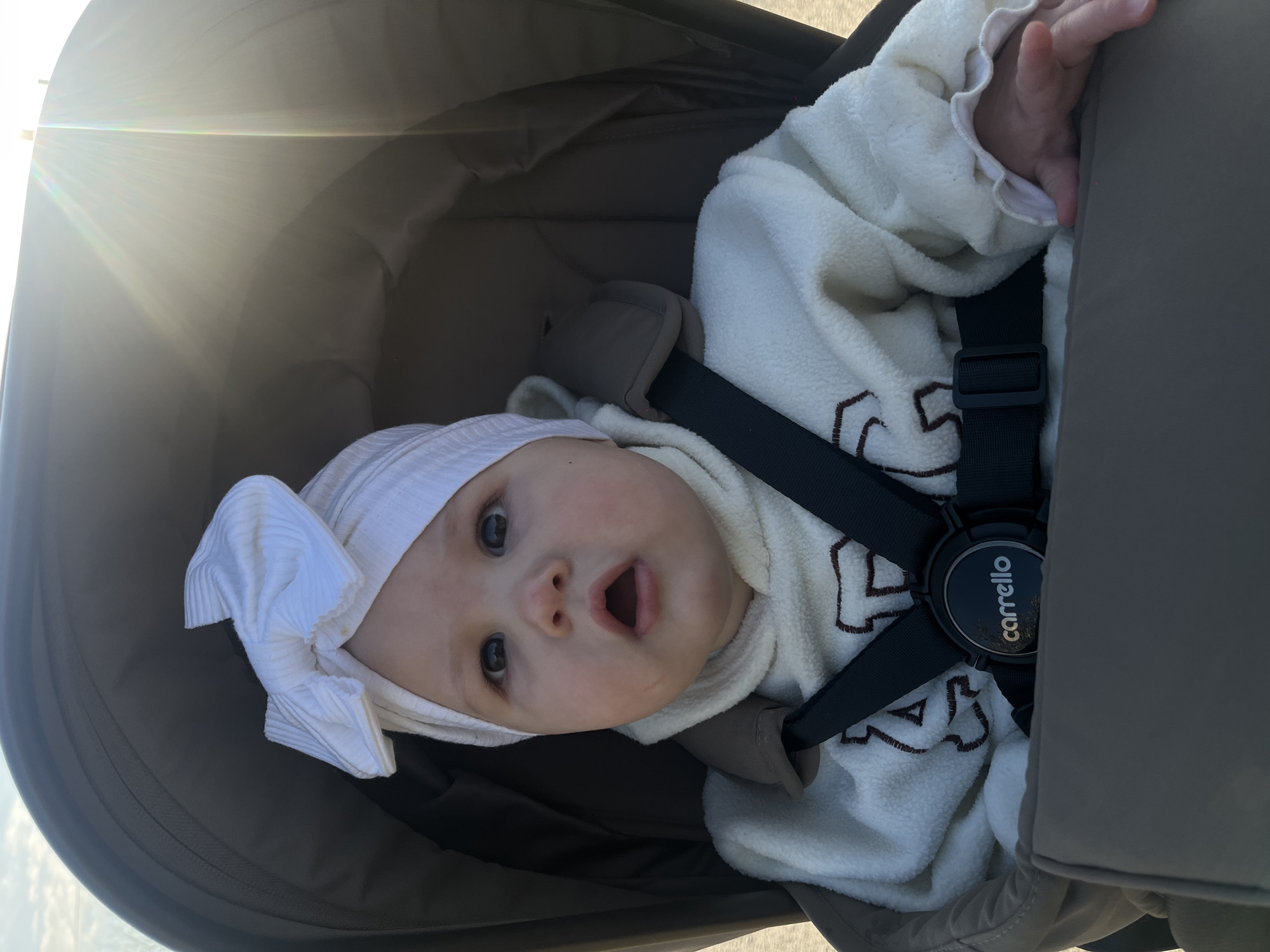 An infant is seen looking upwards from within a dark-colored stroller or car seat, captured in a diagonally oriented, brightly lit photograph. The baby has fair skin, large dark eyes, and an open mouth forming an "O" shape, conveying an expression of surprise or curiosity. A white, ribbed head wrap with a prominent bow on the left side adorns the baby's head. The baby is dressed in a cozy, white fleecy garment featuring brown embroidered patterns.
The stroller or car seat is dark gray or brown and has a visible five-point harness system. A central black buckle on the harness prominently displays the brand name "carrello" in yellow text. Padded shoulder straps are also visible. Bright sunlight streams in from the left side of the frame, creating a strong lens flare and illuminating the scene. The canopy of the stroller provides some shade but allows the sun's rays to enter.
The image depicts a casual outdoor moment, likely during the day, given the strong sunlight and clear weather. The baby appears engaged with the camera, possibly reacting to the photographer. The setting is outdoors in Novopokrovskaya, Russia.