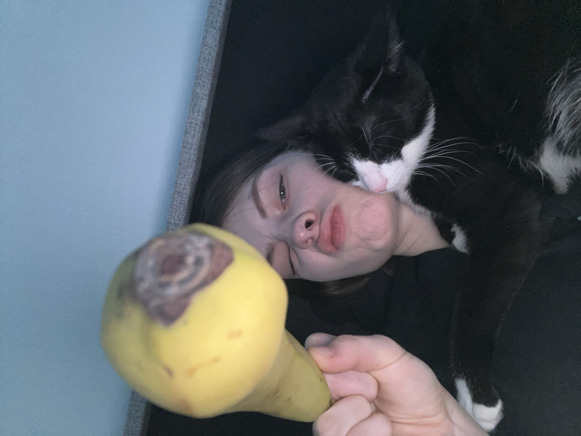 A woman is lying on a dark surface, possibly a couch, with a black and white cat resting on her head and neck. She is holding a yellow pear, which has a brown, rotten spot at the top, in her hand in the foreground. Her face is scrunched up as if she is making a silly expression. A light blue wall can be seen in the background. The location cannot be determined based on the image provided.