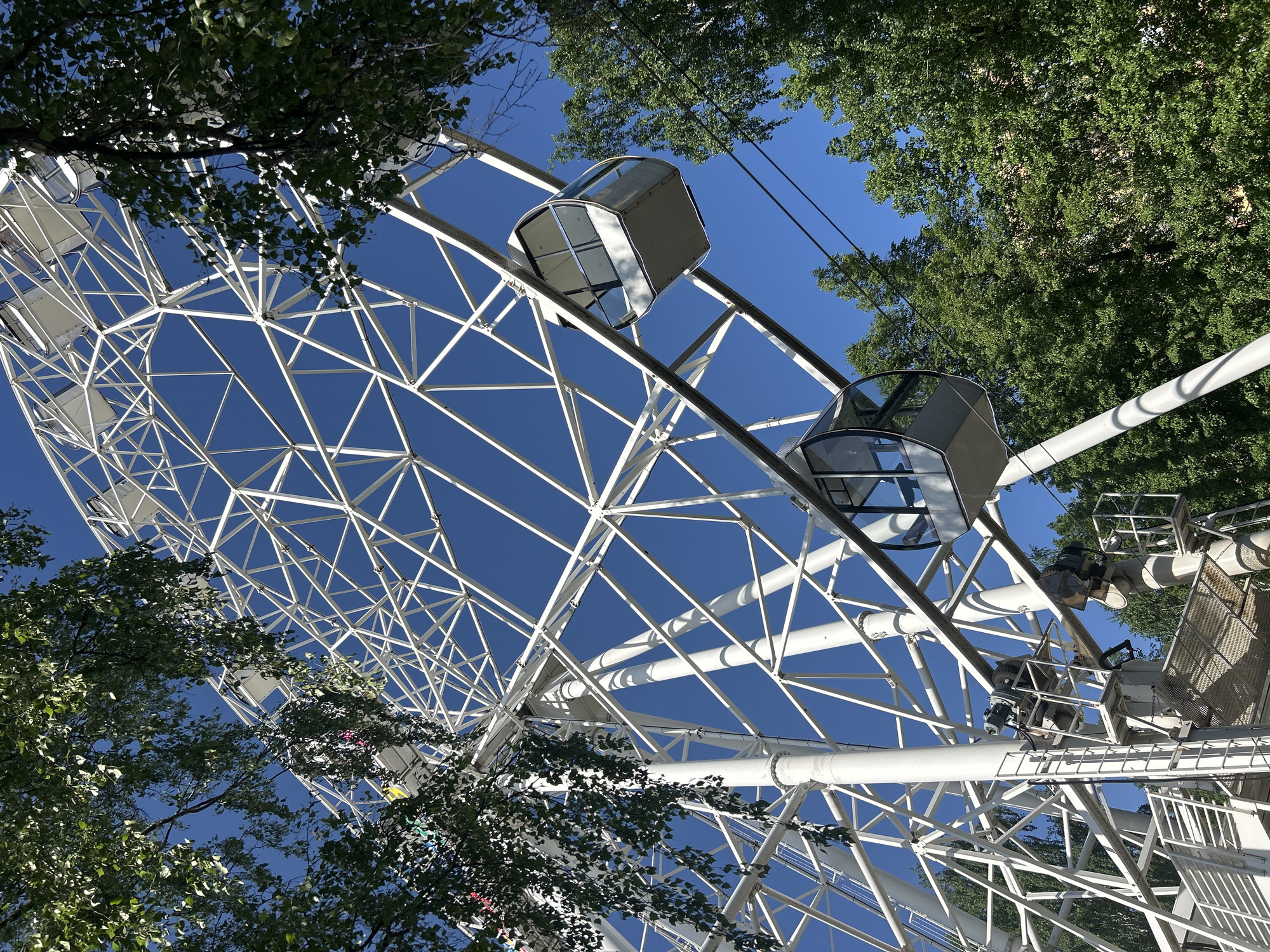 The image shows a Ferris wheel in Izhevsk, Russia, viewed from below. The structure is white, with enclosed passenger cabins. The sky is clear and blue, visible through the wheel's framework. Trees with green leaves surround the wheel, partially obscuring the view. Maintenance platforms are visible on the wheel's structure.