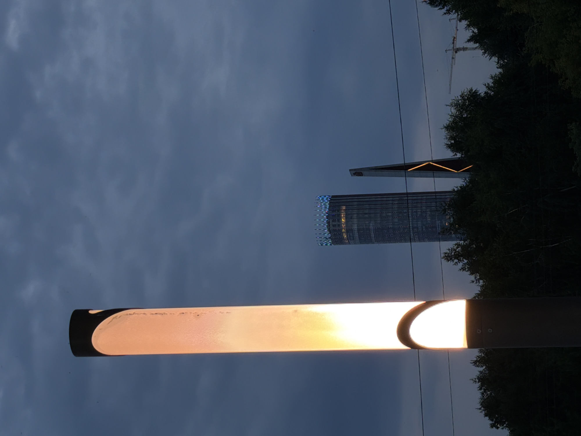 In the twilight of Yekaterinburg, Russia, a tall, cylindrical lamp post stands prominently in the foreground, its upper section illuminated with a warm, orange glow. The light source within creates a diffused luminescence through the translucent material of the post. Behind this, two modern skyscrapers pierce the darkening sky. The taller of the two, on the left, is a dark glass tower adorned with a band of blue and white lights near its apex. To its right, a more angular structure with a sharp, geometric design features a striking orange illuminated line that forms a V-shape. A dense line of dark green trees forms a barrier across the middle ground, with electrical wires running horizontally above them. In the distance, a construction crane is faintly visible against the cloudy, dusk sky.