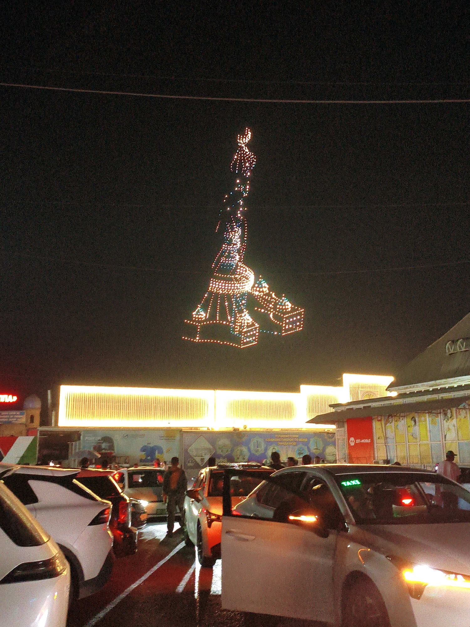 This is a nighttime, medium-angle, eye-level shot of a street scene in Bokhtar, Tajikistan, featuring a large, illuminated Christmas tree-like structure suspended in the air against a dark sky. The structure is made of numerous small lights and has a distinct conical shape with layered embellishments. In the foreground, several cars are stopped on a wet road, reflecting the surrounding lights. A white sedan is prominently featured on the right, with its headlights and hazard lights illuminated. A green "TAXI" sign is visible on its roof. To the left, another white car is partially visible. Behind the cars, a building with bright, yellow, horizontal lighting is visible. People are present, with two individuals standing near the cars on the right and several others further back. The overall atmosphere suggests a festive, possibly holiday-related event in a city setting.