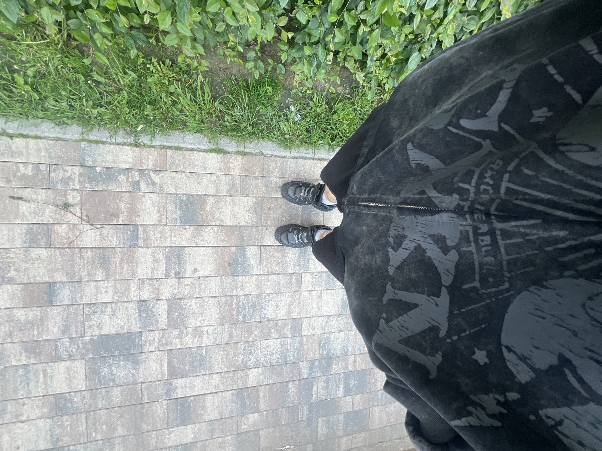 An overhead, first-person perspective photograph captures a person standing on a paved surface next to a strip of green vegetation. The subject's lower body is visible, dressed in dark, possibly black, loose-fitting trousers or sweatpants and a matching dark top or jacket. The clothing features light grey, distressed-looking graphics, including stylized figures, what appears to be a star, and partial text. On the top, letters that resemble "RACKIEL" or "RACKIBLE" are faintly discernible amidst the patterns and fabric folds, along with other fragmented words. The person is wearing dark, chunky athletic or casual shoes with light-colored laces and visible white socks at the ankles.
The setting is outdoors during the daytime, under what appears to be overcast or diffused light conditions, as no strong shadows are cast. The ground beneath the person is composed of rectangular paving stones or bricks, laid in a stretcher bond pattern, showcasing variations in shades of grey, blue, and brown. To the left side of the frame, bordering the paved area, is a vibrant strip of green grass interspersed with some weeds and dirt, backed by dense, leafy green bushes or shrubs. A small piece of white debris is visible in the grass. The overall scene suggests a casual walk or pause in a park, garden, or urban pedestrian area. The location is unknown, Russia.