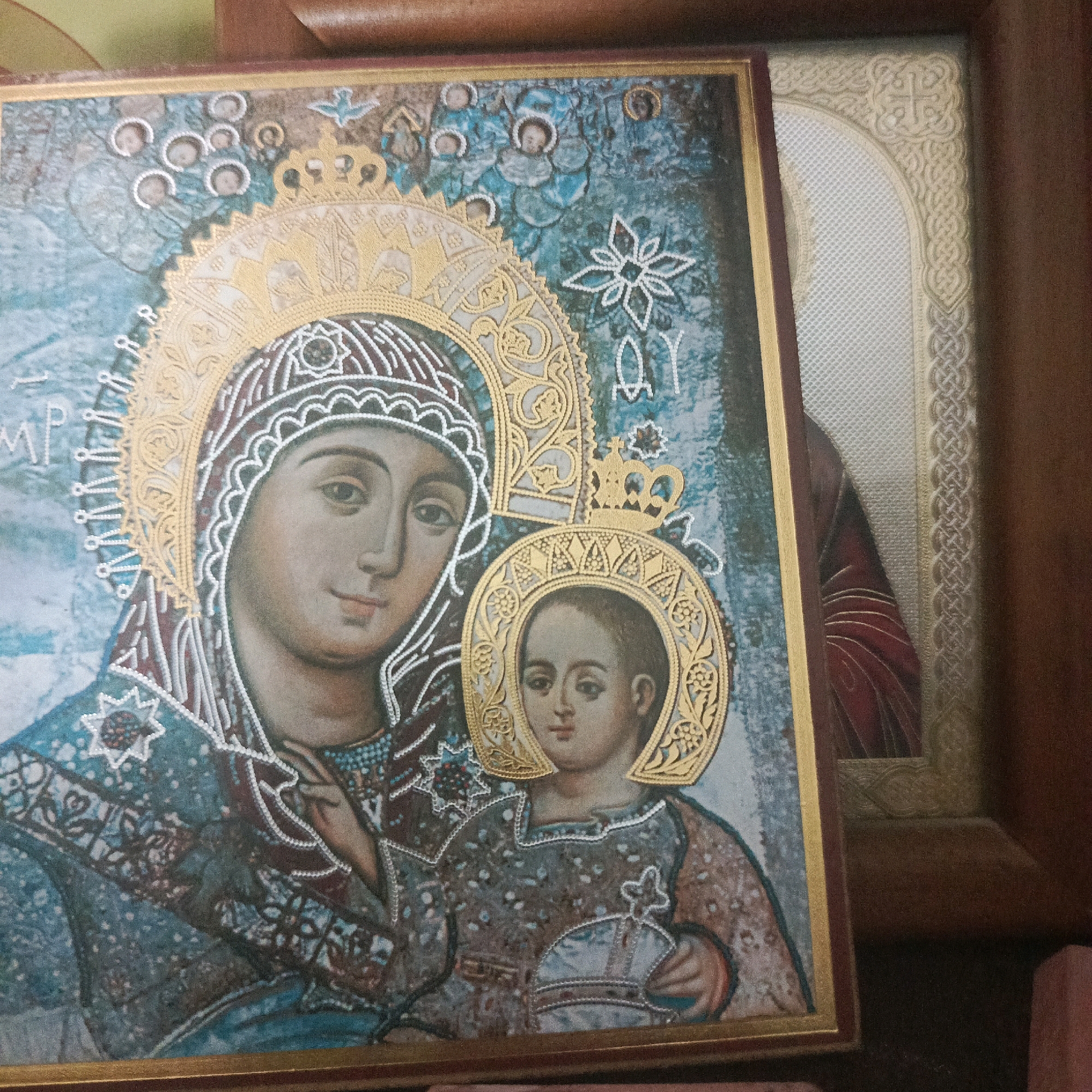 The image shows two framed religious icons. The icon in the foreground is a depiction of the Virgin Mary and the Christ Child. The Virgin Mary wears a blue robe adorned with a golden crown and a detailed halo. The Christ Child is held in her arms, also wearing a robe and a halo. Both figures have serene expressions. The icon is richly decorated with golden ornamentation and appears to be made with various textured materials, possibly including small beads. The background of the icon has a textured, mottled blue and grey appearance with some angelic figures and decorative elements.
To the right and slightly behind the foreground icon, a second framed icon is visible. This icon features an arched design with intricate, interlaced knotwork patterns in a light, textured material. A cross is visible within the upper arch. The visible portion of the image within this second frame is a dark red fabric, possibly a garment.
The icons are placed on a surface that is not fully visible, but appears to be dark. The overall lighting suggests an indoor setting. The provided information indicates the location of this image is in the city of Стрехнино, Russia.