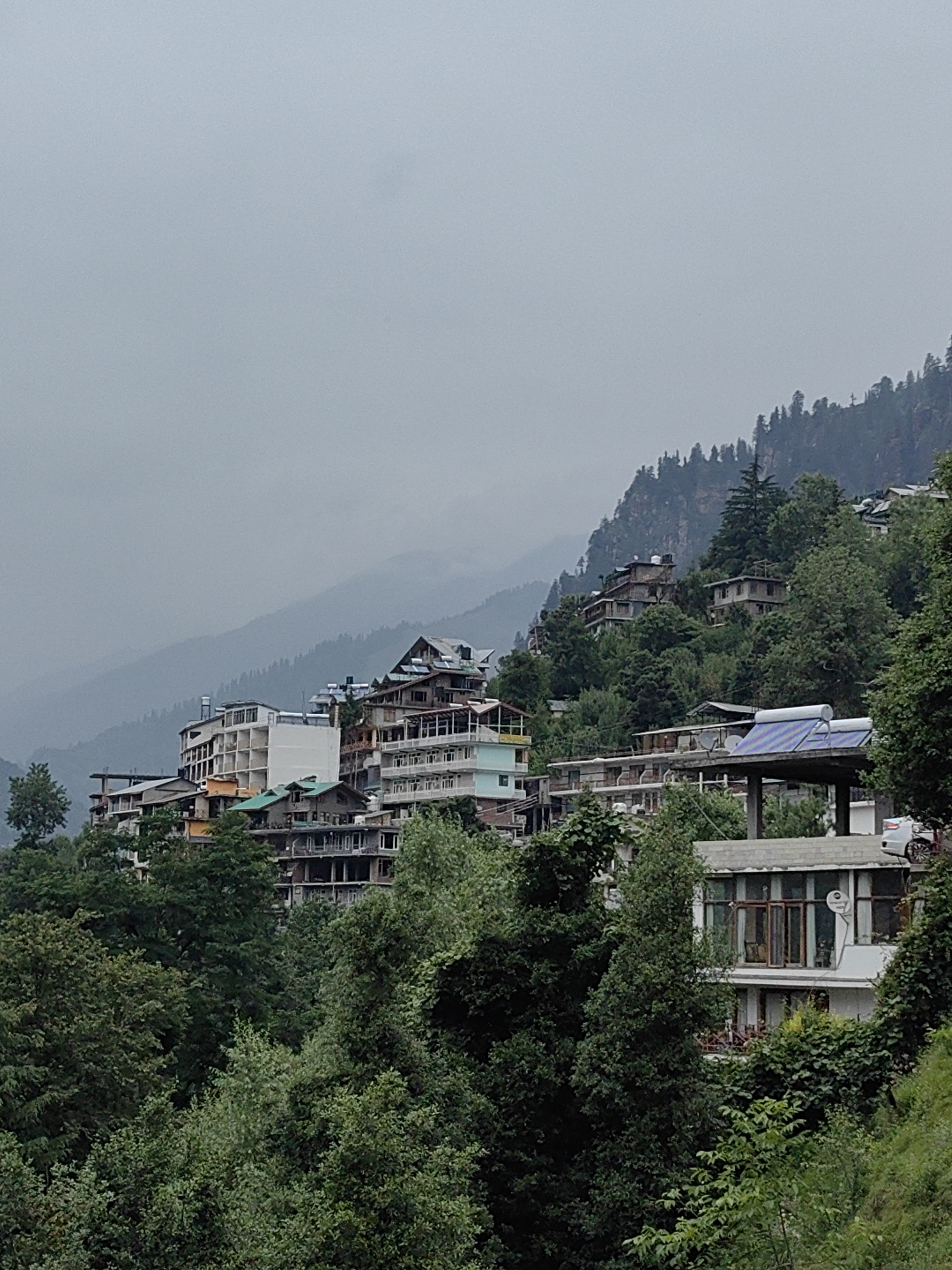 The image depicts a view of a hillside village in Vashisht, India, on an overcast day. The scene is characterized by numerous buildings nestled into the steep terrain, surrounded by dense green foliage. The architecture is varied, with multi-story structures exhibiting a mix of modern and traditional designs, some appearing to be hotels or guesthouses. Many buildings have balconies, and some are adorned with solar water heaters.
The foreground is dominated by lush trees and vegetation, suggesting a natural, mountainous environment. In the midground, the cluster of buildings rises up the hillside. The background shows more layers of misty mountains, shrouded in a hazy, diffused light characteristic of cloudy weather. The overall mood is tranquil, with the muted colors and the overcast sky creating a serene atmosphere. There are no visible people or specific activities taking place, suggesting a general landscape or architectural view of the village. The time of day is difficult to ascertain precisely due to the overcast conditions, but it appears to be daytime. The location cues, such as the architecture and the dense greenery, are consistent with a mountainous region like Vashisht, India. No text is visible in the image.