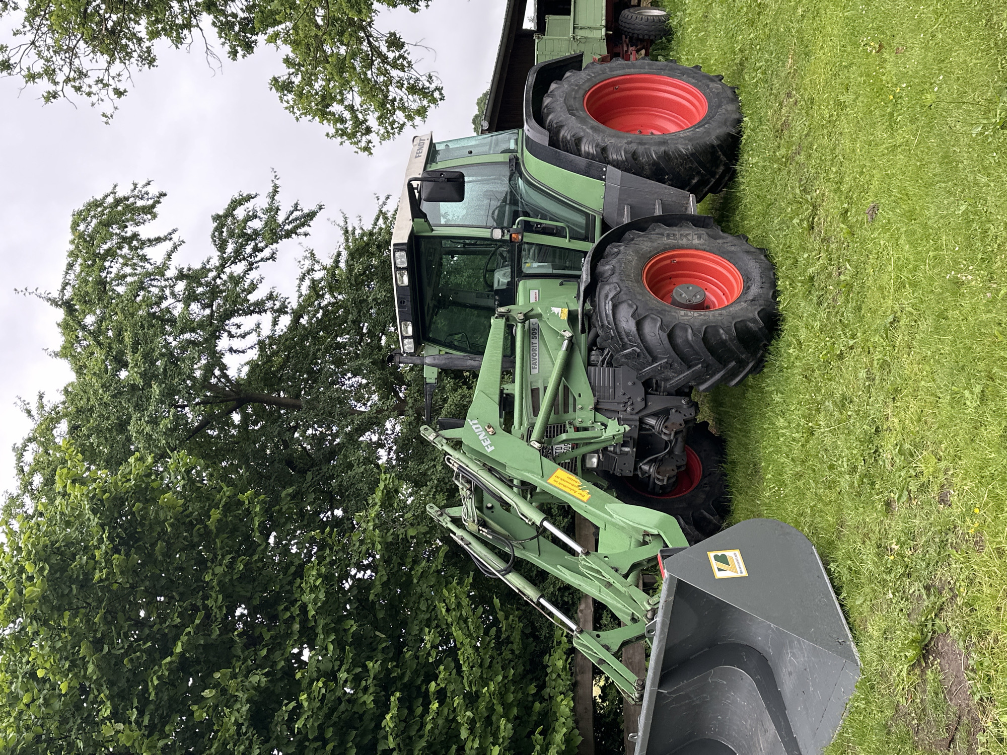This image, captured in Deinste, Germany, depicts a green Fendt Favorit 509 C tractor with a front-end loader and an attached grey bucket, parked on a patch of green grass. The tractor features large black tires with prominent red wheel hubs on both axles. The front-end loader arms are green, with visible silver hydraulic cylinders. A yellow rectangular sticker with German text is affixed to the right loader arm. The grey bucket displays a yellow and black "ZB" logo with the text "BRENNER UND LACKE". The tractor's cab has multiple roof-mounted lights and a rearview mirror on the left side. The "FENDT" brand name is visible on the upper right side of the cab. Behind the tractor, a portion of a green, two-axle trailer with white wheel hubs is visible. The background consists predominantly of lush green deciduous trees, which frame the upper and left portions of the image. The sky above is overcast and light grey. No individuals are present in the scene.