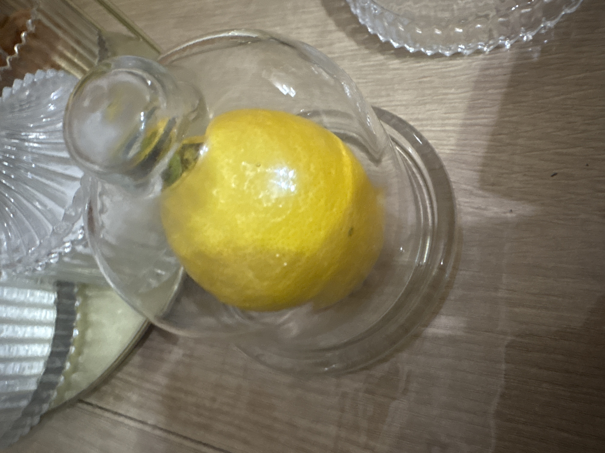 A top-down view reveals a lemon resting in a clear glass container with a matching lid. The scene is set on a wooden surface, with other pieces of glass decor present in the frame.
