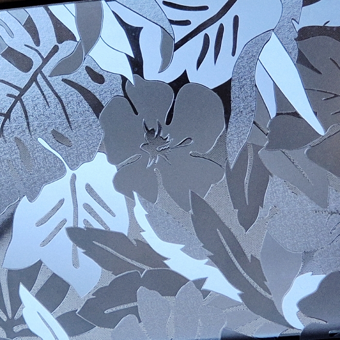 This scene, located in Strasbourg, France, presents a close-up view of a static, decorative surface. The surface displays a monochromatic botanical pattern rendered in various shades of gray, silver, and off-white. The pattern features an overlapping arrangement of stylized leaves and flowers. Prominent leaf shapes include broad, palmate leaves reminiscent of Monstera, elongated leaves with serrated edges, and simpler oval-shaped foliage. A distinct five-petal flower with detailed stamen is centrally visible among the leaves. The texture of the pattern suggests a frosted or etched glass finish, or a similar opaque material, with lighter areas appearing reflective and darker areas providing contrast. There are no human subjects, active interactions, or discernible environmental elements beyond the patterned surface itself.