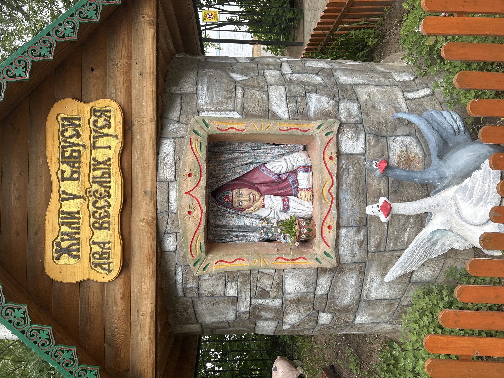 The image depicts an outdoor scene featuring a whimsical, fairy-tale-themed structure. At the center is a stone-like cylindrical building with a wooden roof. Above the window of the building, a wooden sign with yellow lettering reads "Жили у бабуси два весёлых гуся" (Once upon a time, there lived two merry geese with grandma). A sculpted figure of a woman in traditional Russian attire, wearing a headscarf, is visible through the window, resting her chin on her hand and looking out. In front of the structure, two large, stylized goose sculptures are positioned, one white and one gray. A small orange wooden fence runs across the foreground. To the left of the main structure, a pink pig sculpture is partially visible. In the background, there is foliage, a body of water (likely the Kama River), and a yellow sign with a warning symbol. This structure is located in the city of Chaikovsky, Russia, specifically in the central park area along the Kama River embankment, known for its recreational and artistic installations.