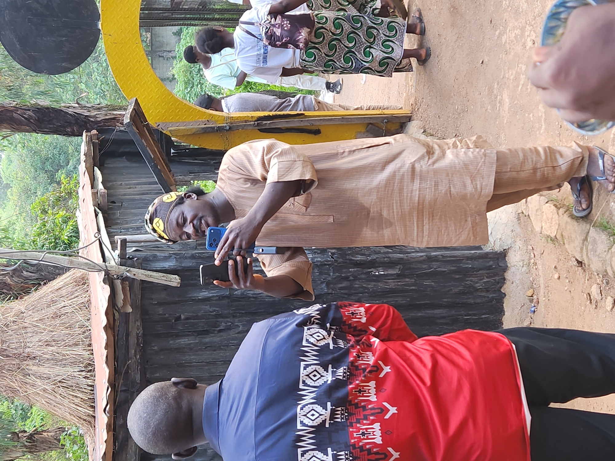 The image depicts an outdoor scene, likely in Jos, Nigeria. In the foreground, a person wearing a blue and red shirt with an intricate white and red pattern is facing away from the camera. To their right, another individual, dressed in a light brown traditional outfit and a patterned headwrap, is holding two smartphones, one in each hand, appearing to film or photograph something. Behind these two figures, a rustic structure with a thatched roof and corrugated metal siding is visible. Further in the background, a yellow archway frames a path leading to other individuals. One person is wearing a white t-shirt with text and a patterned skirt, carrying a backpack. The environment appears somewhat overgrown with vegetation, and the ground is dirt. The lighting suggests it is daytime.