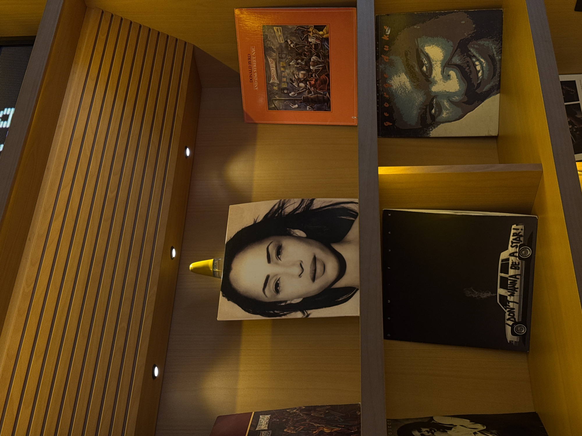 The image displays a warm-toned, wooden display shelf, likely indoors in Yerevan, Armenia, showcasing several vinyl record covers. The shelves are made of light-colored wood, with some sections featuring vertical wooden slats. Small, recessed spotlights are embedded within the shelving, casting a warm glow on the items.
On the top shelf, two record covers are visible. The left cover is predominantly orange, featuring a detailed illustration of a street scene with numerous figures. Prominently displayed text on this cover reads "DONALD BYRD AND 125th STREET, N.Y.C." and "DONALD BYRD." To its right, another album cover features a stylized, close-up illustration of a person's face in shades of green, blue, and black.
On the middle shelf, two additional record covers are displayed. On the left, a black and white album cover presents a portrait of a woman's face, with a small, yellow, conical object resting on top of the cover. To its right, a black album cover showcases a white line drawing of a station wagon with exhaust smoke, and the text "I DON'T WANNA BE A STAR!" clearly visible on the car's side. The letters "D O M F U N K" are partially visible along the left edge of this album.
No human activity is depicted; the scene is a static arrangement of the album art. The overall impression is that of a curated collection or display in a well-lit interior.