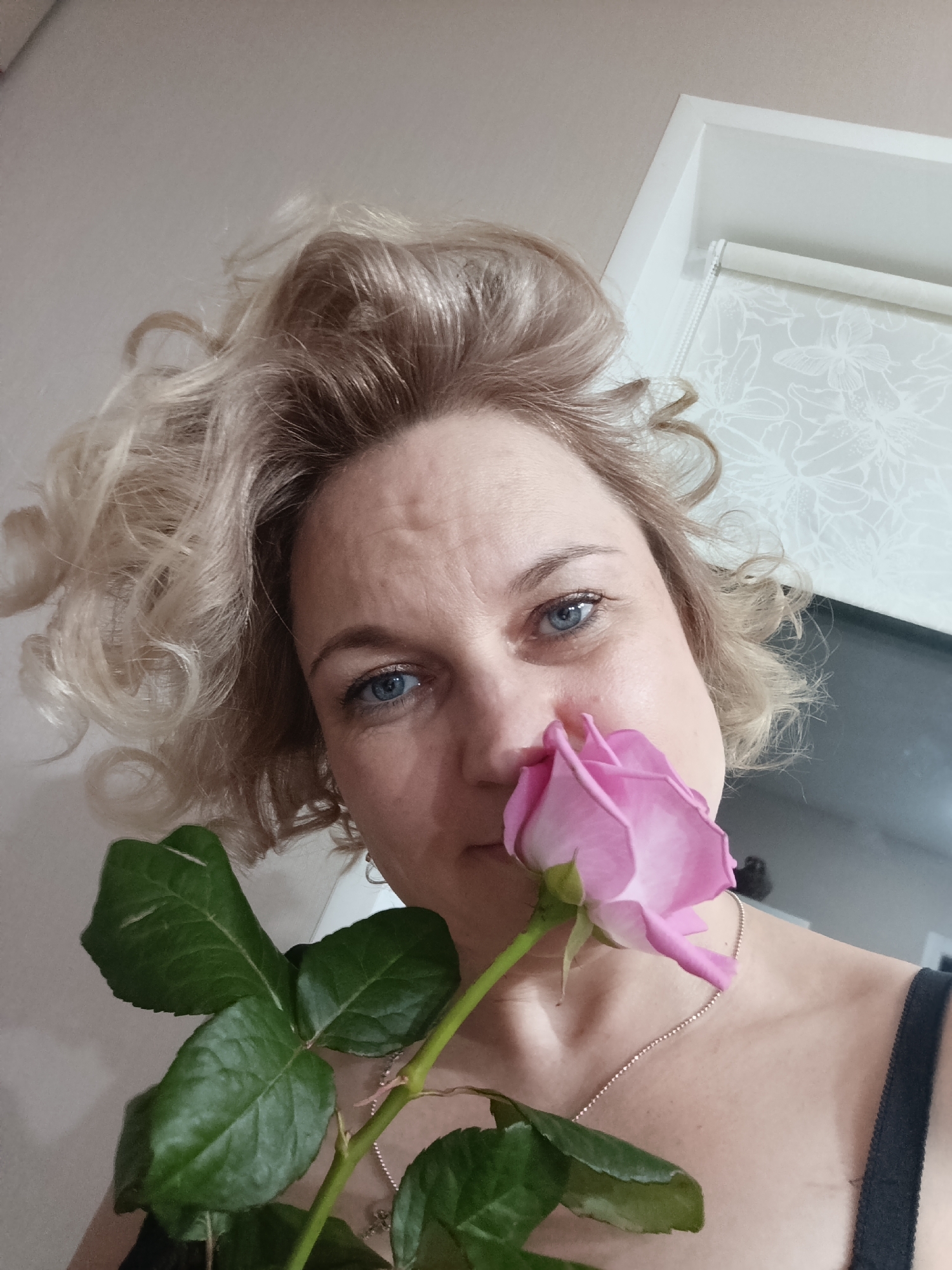 The image displays a close-up, low-angle indoor portrait of an adult female, reportedly taken in the city of Марковский, Russia. The subject, possessing fair skin, blue eyes, and voluminous blonde curly hair, is positioned centrally, gazing directly at the camera. She holds a single pink rose with an intact green stem and leaves; the bloom is situated near her nose, indicating the action of smelling. A dark garment strap is visible on her right shoulder, and a thin, light-colored beaded necklace is worn around her neck. The background consists of a light-colored wall and ceiling. In the upper right quadrant, a white architectural frame is visible, behind which a light-colored roller blind adorned with a subtle white floral pattern is partially discernable.