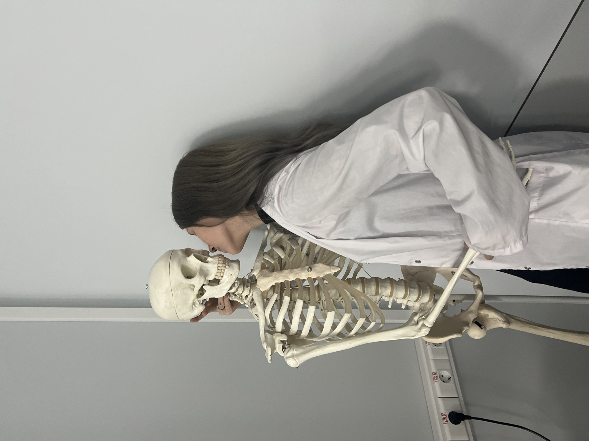 A female individual, viewed in profile, is depicted indoors interacting with a full-body anatomical skeleton model. She has long, wavy, medium-brown hair and is wearing a white lab coat over a dark garment. Her lips are pursed, touching the left temporal region of the skeleton's skull, while her right hand gently supports the posterior neck/skull area of the model. A ring is visible on her right middle finger. The skeleton, an off-white plastic anatomical model with visible metallic joint articulation pins, stands upright. The background consists of light gray walls. On the left, a white electrical conduit or power strip is mounted horizontally on the wall, featuring multiple "220 B" electrical outlets, one of which is occupied by a black plug and cable. The "220 B" marking on the outlets is consistent with electrical standards found in Moscow, Russia. The setting appears to be a classroom, laboratory, or medical education facility.