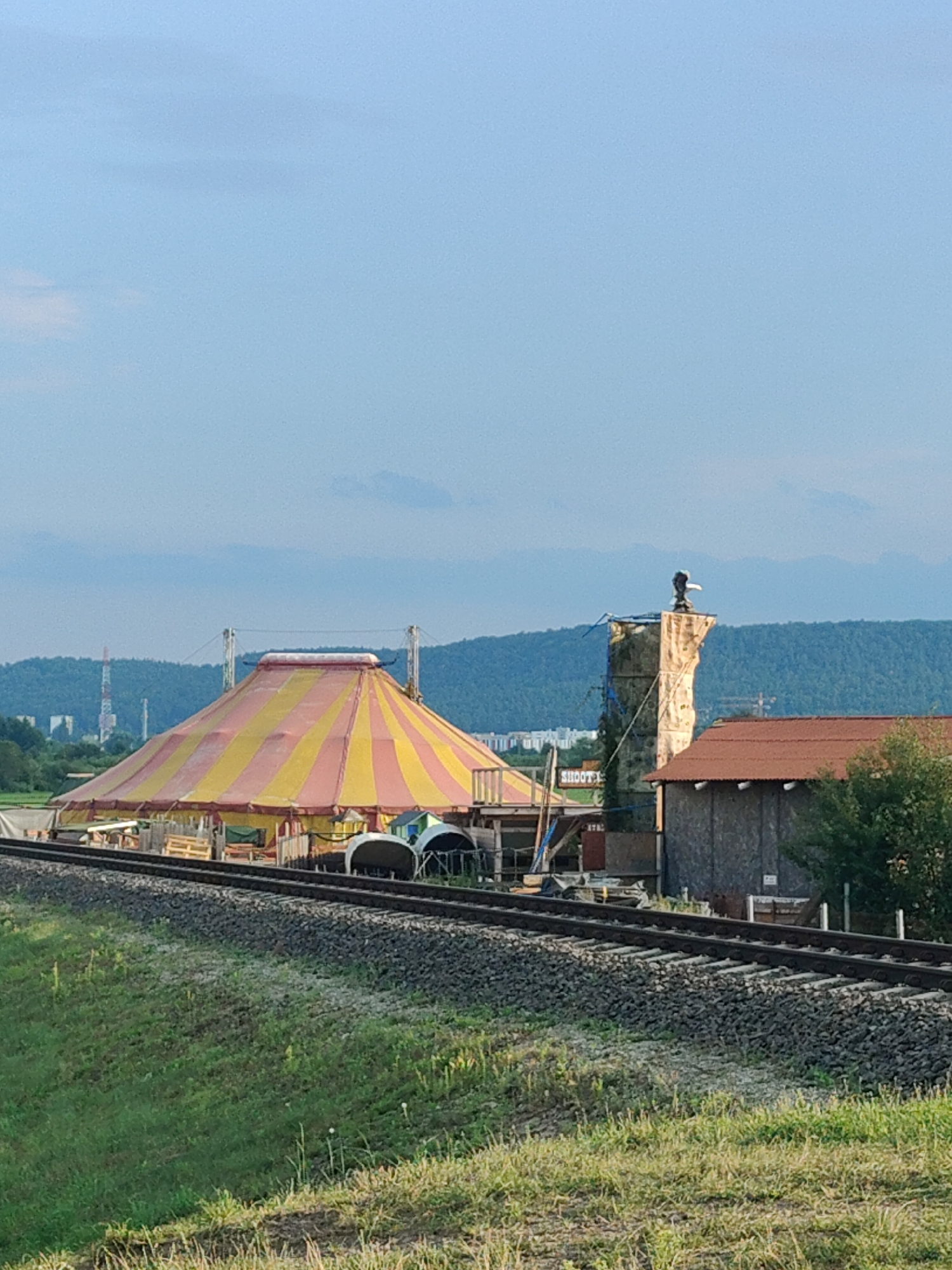This is a full-frame, eye-level, vertical shot of a circus tent and a climbing wall structure in Dębogórze, Poland, likely during daytime under a clear sky with some clouds on the horizon. The foreground is dominated by a railway track running diagonally from the bottom left to the top right. The ground slopes upwards from the tracks, covered in green grass with some sparse weeds and dry patches.
The main subject on the left is a large, traditional circus tent with a striped pattern of red and yellow. It appears to be set up, with poles and ropes visible around its perimeter. Behind the tent, in the middle ground, there's a tall, rough-textured, stone-like structure that resembles a climbing wall. A figure, possibly a person in dark clothing, is perched at the very top of this structure, holding what appears to be a staff or a spear.
To the right of the climbing wall, there is a small, brown, wooden building with a reddish-brown roof. Further to the right and slightly behind this building, there is a cluster of trees. In the background, a range of hazy, green hills and mountains stretches across the horizon. The sky above is a pale blue, with some wispy clouds. In the far left background, industrial structures, possibly towers or chimneys, are visible against the distant hills. The overall scene suggests an outdoor event or a temporary setup.