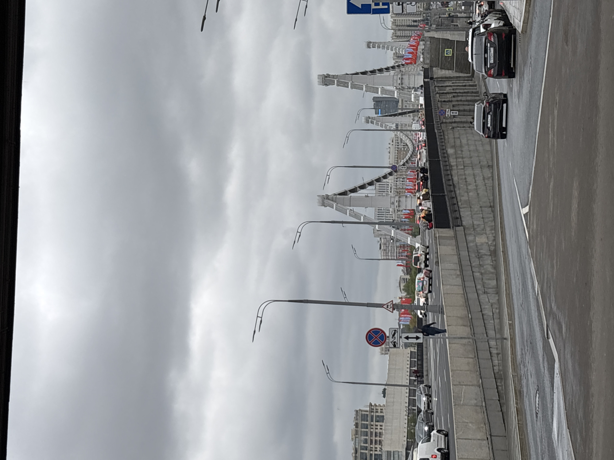 The image shows a view of the Krymsky Bridge in Moscow, Russia. The bridge is visible with its distinctive white arches. The sky is overcast. Traffic is present on the roadway beside the bridge. Street lights line the bridge. Red flags are visible on the bridge. Buildings are visible in the background.