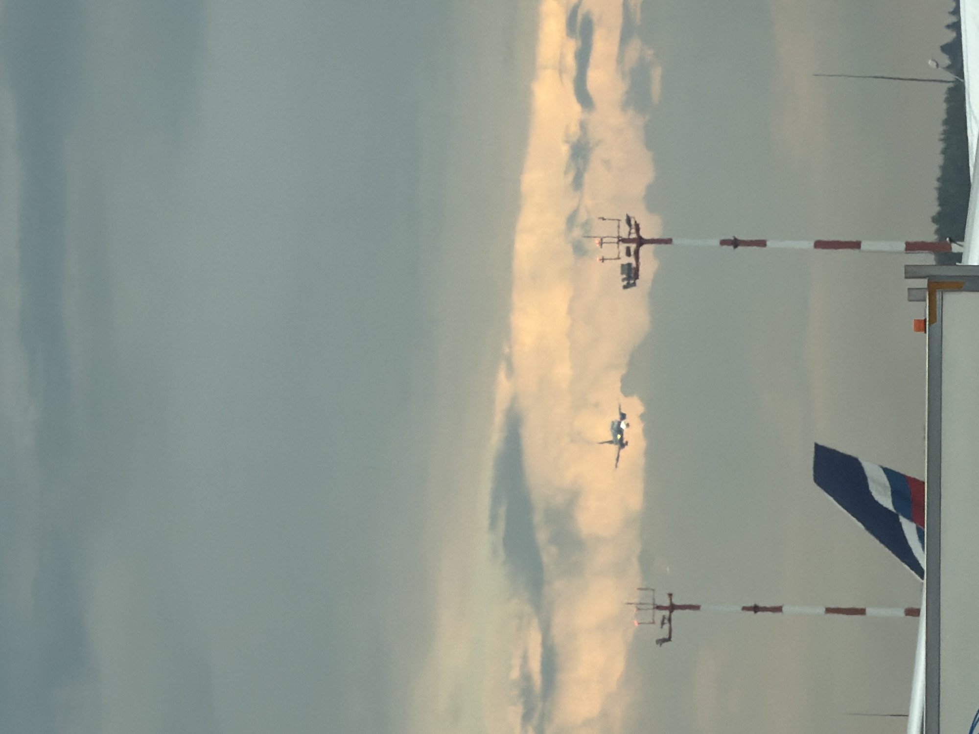 The image, captured in Khimki, Russia, shows an overcast sky with streaks of golden clouds near the horizon. In the middle ground, a jet airplane is visible in flight against the cloudy sky. In the foreground, to the left, the tail fin of a stationary airplane with a blue, white, and red design is partly visible. Two airport navigation lights, one on the left and one on the right, are also present. The navigation light on the left is a pole with red and white bands, topped with red lights. The navigation light on the right is a taller pole with a similar red and white banding and lights at its top. The overall scene suggests an airport environment under a dramatic sky.