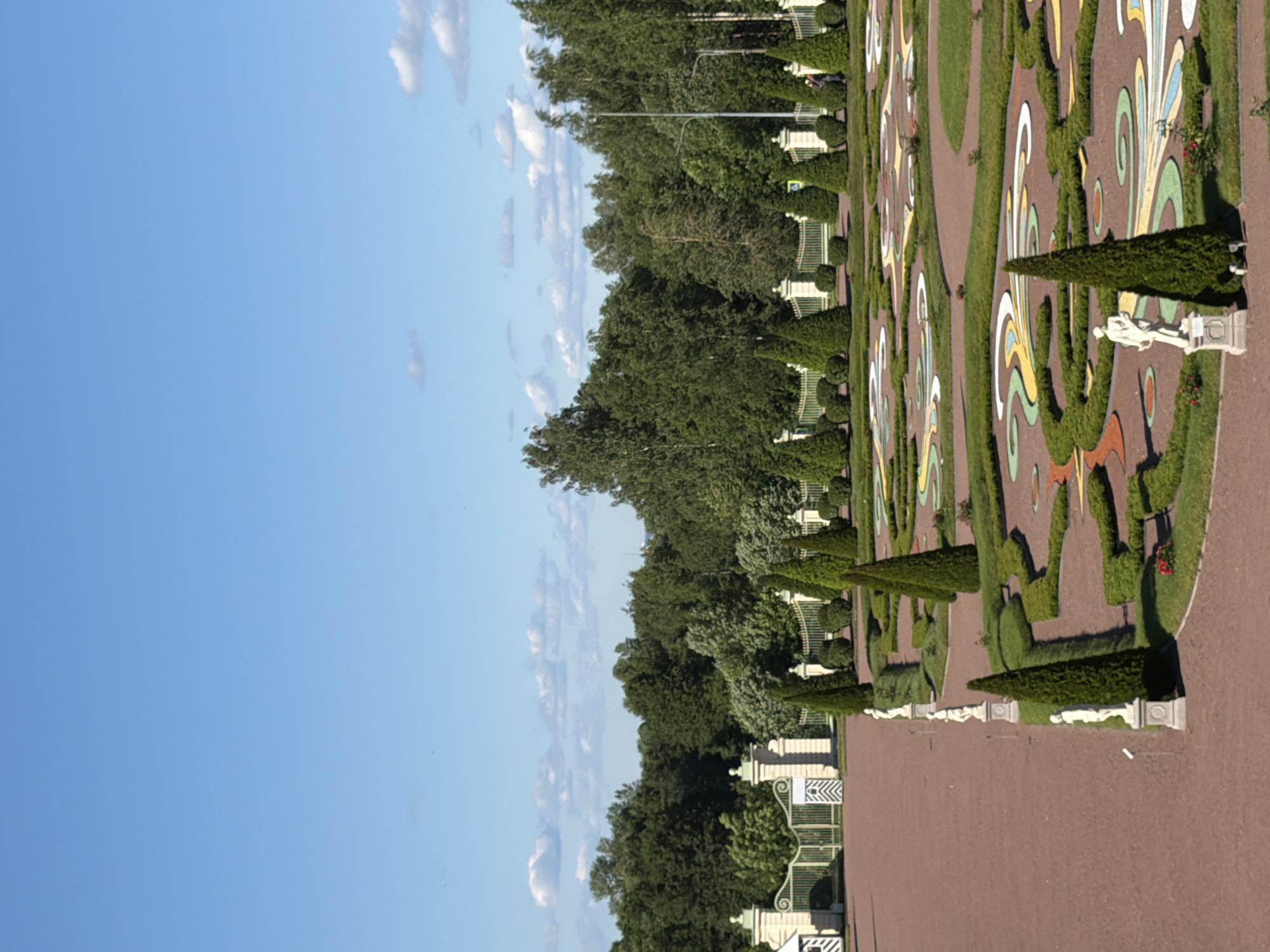 This is a vertical, eye-level, full shot of a formal garden with a patterned landscape and trees under a clear blue sky with a few scattered white clouds. In the foreground, a reddish-brown gravel path leads to a meticulously manicured garden bed. This garden bed features a colorful, intricate pattern of greenery, orange, yellow, white, and light blue, laid out in swirling shapes. Several conical evergreen trees are interspersed throughout the garden, along with a few white statues of robed figures standing on pedestals. To the left, a row of three similar statues lines the gravel path. Beyond the garden bed, a golden metal fence with white pillars and decorative elements stretches across the scene. Behind the fence, a dense line of tall, green trees forms a backdrop. In the far distance, more trees and foliage are visible, and a few distant buildings can be seen through the trees. The sky is a bright, clear blue, with a few small, wispy white clouds in the upper portion of the frame. The overall impression is one of order, beauty, and grand scale.