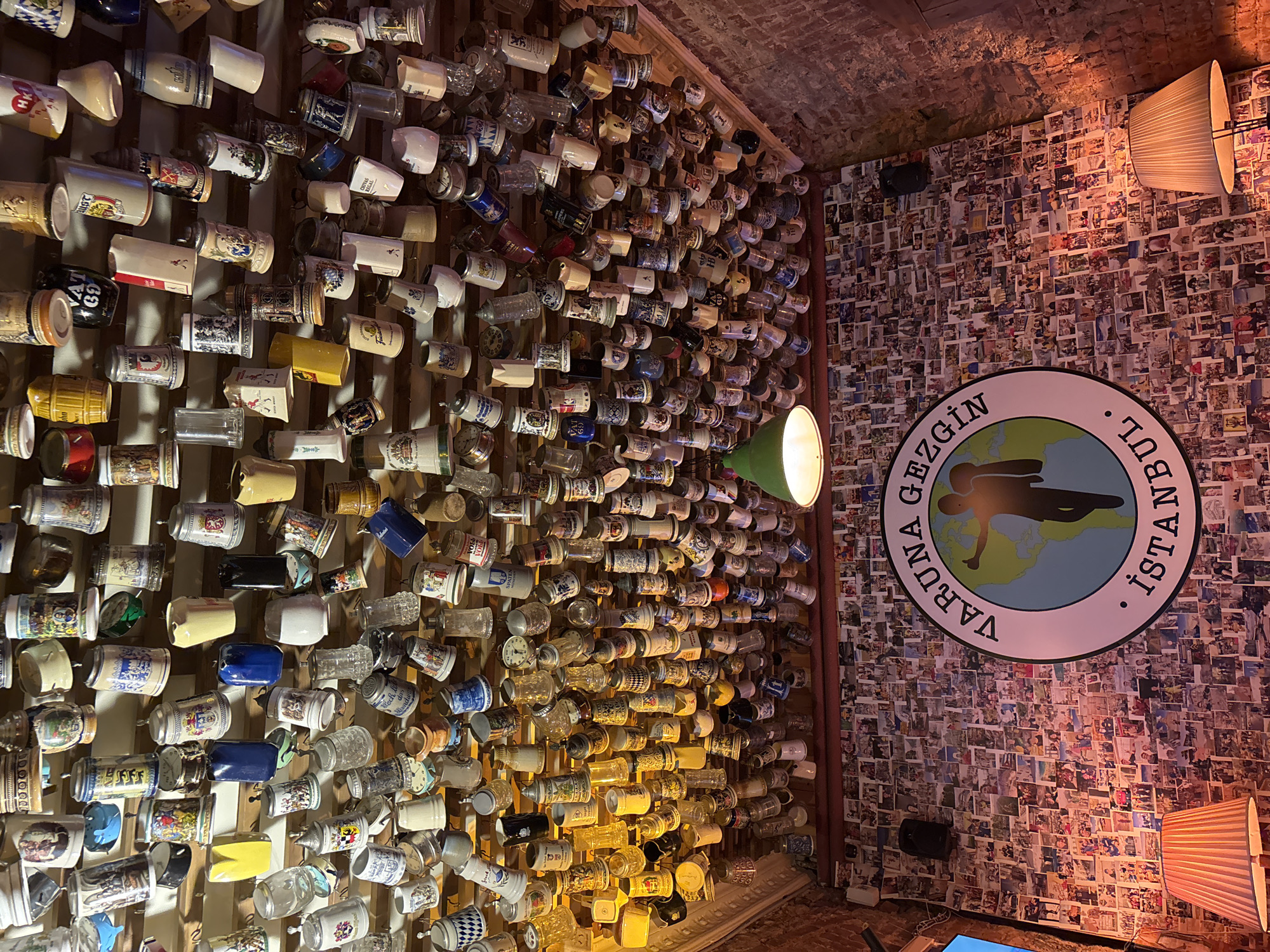 The image shows an interior scene of a dimly lit establishment, possibly a bar or restaurant, adorned with a vast collection of beer steins and mugs displayed on wooden shelves covering one wall. The steins vary in size, color, and design, with many featuring intricate patterns and painted images, suggesting a theme related to German or European culture. Another wall is entirely covered with a collage of photographs, creating a textured and nostalgic ambiance.
Dominating the photo collage wall is a large circular sign with text reading "VARUNA GEZİGİN İSTANBUL" (Varuna Travel Istanbul). The sign contains an illustration of a globe with a silhouette of a person jumping or dancing. A green lamp with a conical shade is visible, casting a warm light on the scene. The ceiling appears to be made of exposed brick. The overall impression is one of a vibrant, perhaps tourist-oriented, venue with a strong sense of character and a rich history, likely located in Beyoğlu, Turkey, as indicated by the text on the sign. The lighting and the abundance of displayed items suggest the establishment is open for business, and the atmosphere appears lively and engaging.