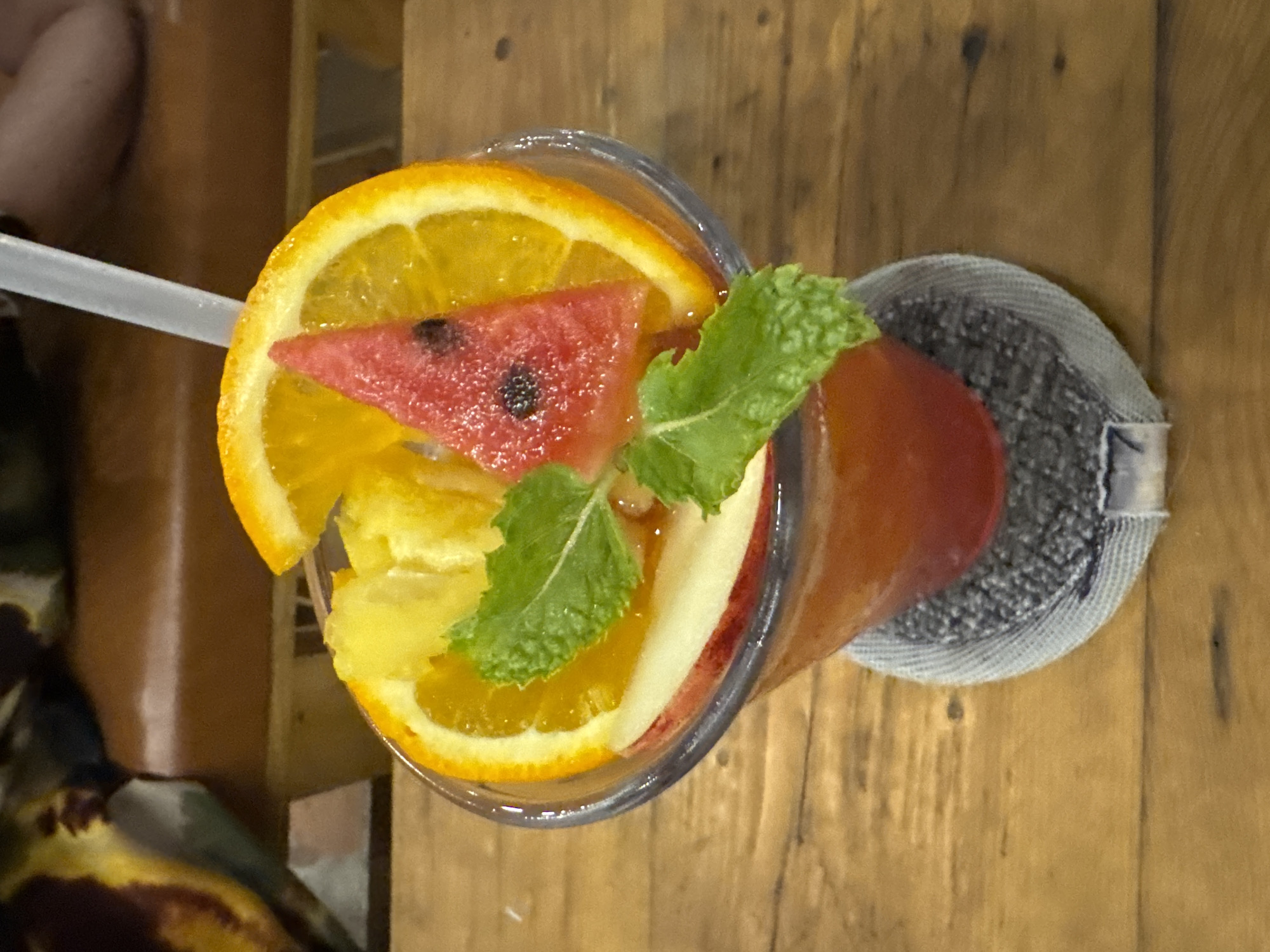 A close-up, overhead shot shows a glass of red juice with fruit garnishes on a wooden surface. The juice is in a clear, transparent glass, and the drink itself is a vibrant red color. Atop the glass are slices of orange and watermelon, a wedge of pineapple, and a sprig of mint. A thin, white straw is inserted into the drink. The glass is placed on a circular, grey coaster. The background is blurred, but appears to be a restaurant or cafe setting, with a leather-upholstered bench and wooden furniture visible. The image is taken from a high angle, focusing on the drink. The overall impression is one of a refreshing beverage in a casual dining environment. The precise location within Bac Nha Trang Ward, Vietnam, is not specified in the image.