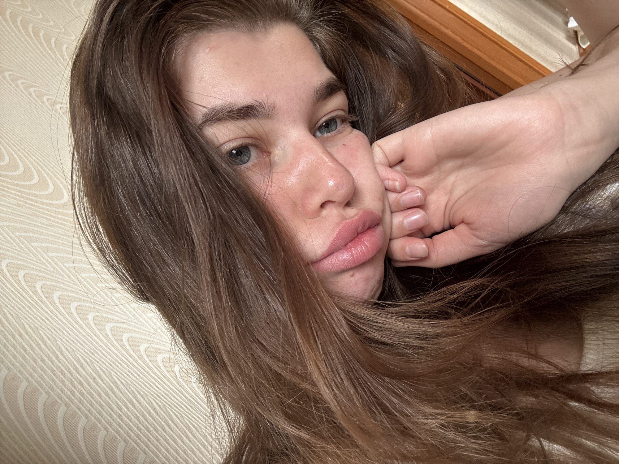 The image shows a close-up, overhead shot of a young woman resting her head on her hand. She has long, brown, wavy hair that obscures much of her face. Her eyes are open and looking to the right, and her lips are slightly parted. A hand, with fingernails that appear to be unpolished or lightly nude, is resting on her cheek, supporting her head. The background consists of a textured, cream-colored wall with a subtle pattern, and a sliver of a wooden frame is visible on the right side of the image. The lighting suggests an indoor setting, possibly a bedroom. There is no indication of the city of Мещёры, Russia in the image itself.