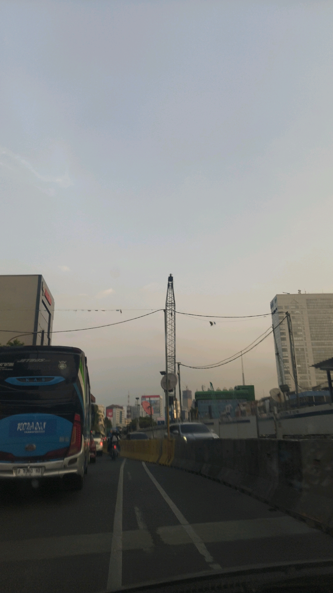 The image captures a view of traffic congestion on a street in Central Jakarta, Indonesia, with a crane visible in the background, indicating ongoing construction. The perspective is from inside a vehicle, looking forward. A blue and black bus with "PUTRA DWI" written on its side is prominent on the left. Multiple cars and a motorcycle are in traffic. Yellow barriers line the right side of the road, separating lanes. In the distance, several high-rise buildings are present, including a white building on the far right. The sky is hazy and appears to be at dusk or dawn. Electrical wires crisscross the scene above the road.