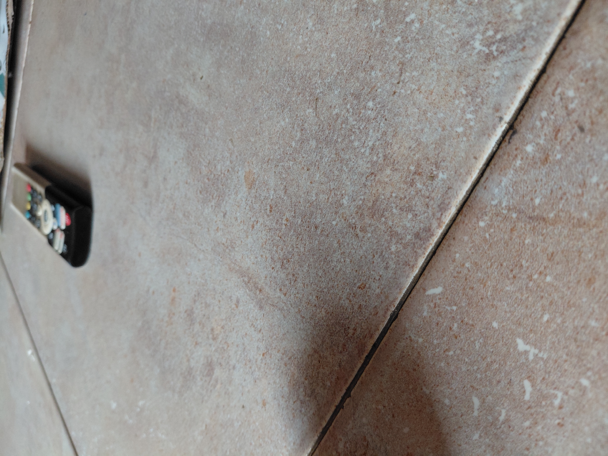 The media file presents a close-up, angled perspective of a tiled surface, likely a floor, within an interior setting. The location is Santa Ana, Venezuela.
The main subjects are the ceramic tiles, which are predominantly a warm brownish-beige. They feature a speckled, textured pattern composed of numerous small white, light brown, and darker brown flecks, creating a natural stone or rustic aesthetic. A dark grout line runs diagonally across the image, clearly separating two tile sections and showing some minor irregularities or debris.
On the left side of the frame, partially visible and slightly out of focus, rests a remote control. It is white and black with a cluster of small, multi-colored buttons, including discernable red, green, blue, white, black, and grey hues.
There is no activity depicted; the remote control is stationary on the tiled surface. The lighting appears even and diffused, characteristic of an indoor environment. No specific time of day or weather conditions can be inferred. No legible text is visible on the remote control or the tiles. The image emphasizes the detailed textures and patterns of the everyday surfaces.