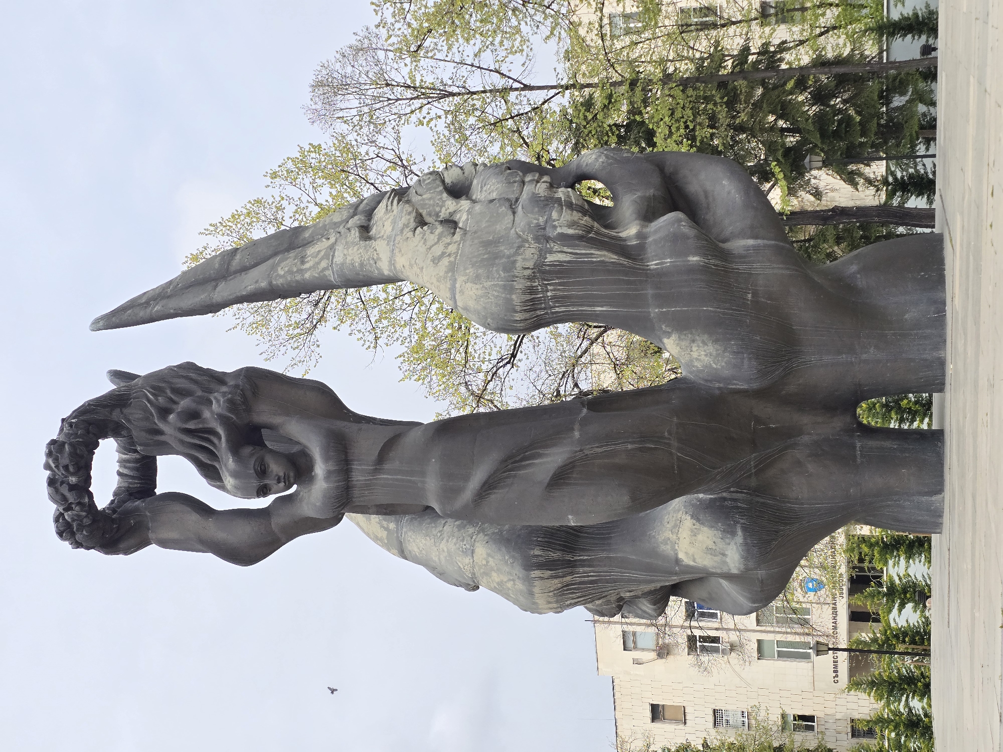 The image shows a bronze sculpture in the city of Unknown, Unknown. The sculpture appears to depict a human figure in a dynamic pose, holding a wreath above its head. The figure is positioned in front of a background that includes trees and a light-colored building. The sculpture is weathered, showing streaks of age and exposure. The sky is overcast.