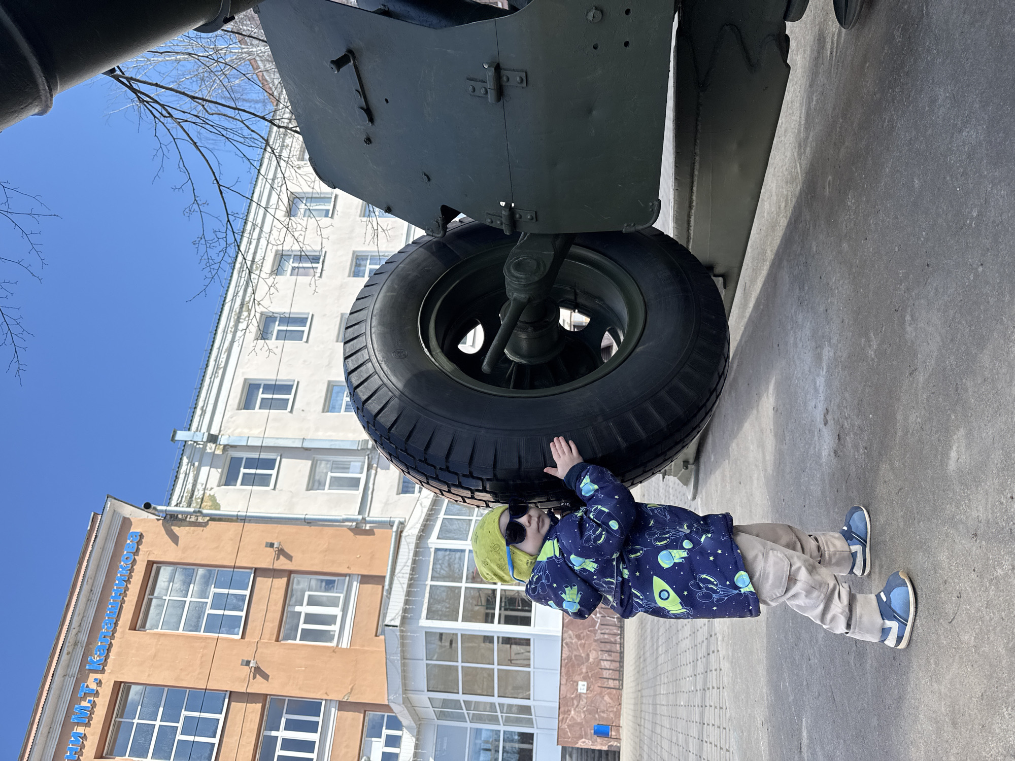 A low-angle shot depicts a young child in sunglasses, a space-themed blue jacket, a light green hat, and beige pants, standing near a large tire of a military vehicle. The child has one hand touching the tire. In the background, there are two buildings. One is a light brown building with white window frames, and blue Cyrillic letters are visible on its side. The other building is white with multiple windows. The scene takes place on a paved surface under a clear blue sky with some bare tree branches visible. The location is Izhevsk, Russia.
