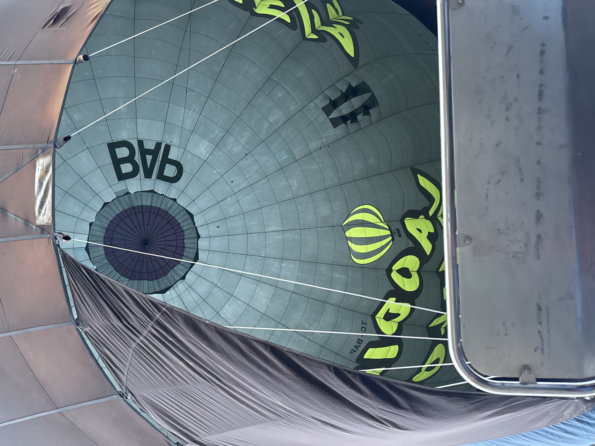 The image provides an internal, upward-looking view within a hot air balloon's envelope, suggesting a moment during its inflation or deflation. The primary subject is the vast, segmented fabric of the balloon, which displays a cool palette of gray and teal. White ropes stretch across the frame, structurally connecting different sections of the envelope.
At the bottom-center of the balloon's dome, a dark purple and black circular vent is visible, contrasting with the surrounding fabric. A section of dark, draped fabric, possibly the skirt or a deflated portion of the balloon, occupies the lower left of the frame. To the right, a metallic structure with a handle and rivets, likely part of the hot air balloon's basket or gondola, frames the scene.
The scene is illuminated by bright daylight, indicating a daytime event. The overall impression is one of anticipation or completion, with the structured lines of the balloon and its materials ready for flight or being prepared for storage.
Prominent black, sans-serif letters spelling "BVB" are clearly visible on the gray-teal fabric. Towards the bottom right, inverted and mirrored characters appear as "TO-BVB" (when viewed normally from below, this would be "BVB-OT"). This inverted text suggests it may be part of an aircraft registration number. Adding to the visual interest are vibrant neon green and yellow stylized motifs, including a distinctive striped hot air balloon icon and abstract, leaf-like designs. The location context provided is Yeşilhisar, Turkey.