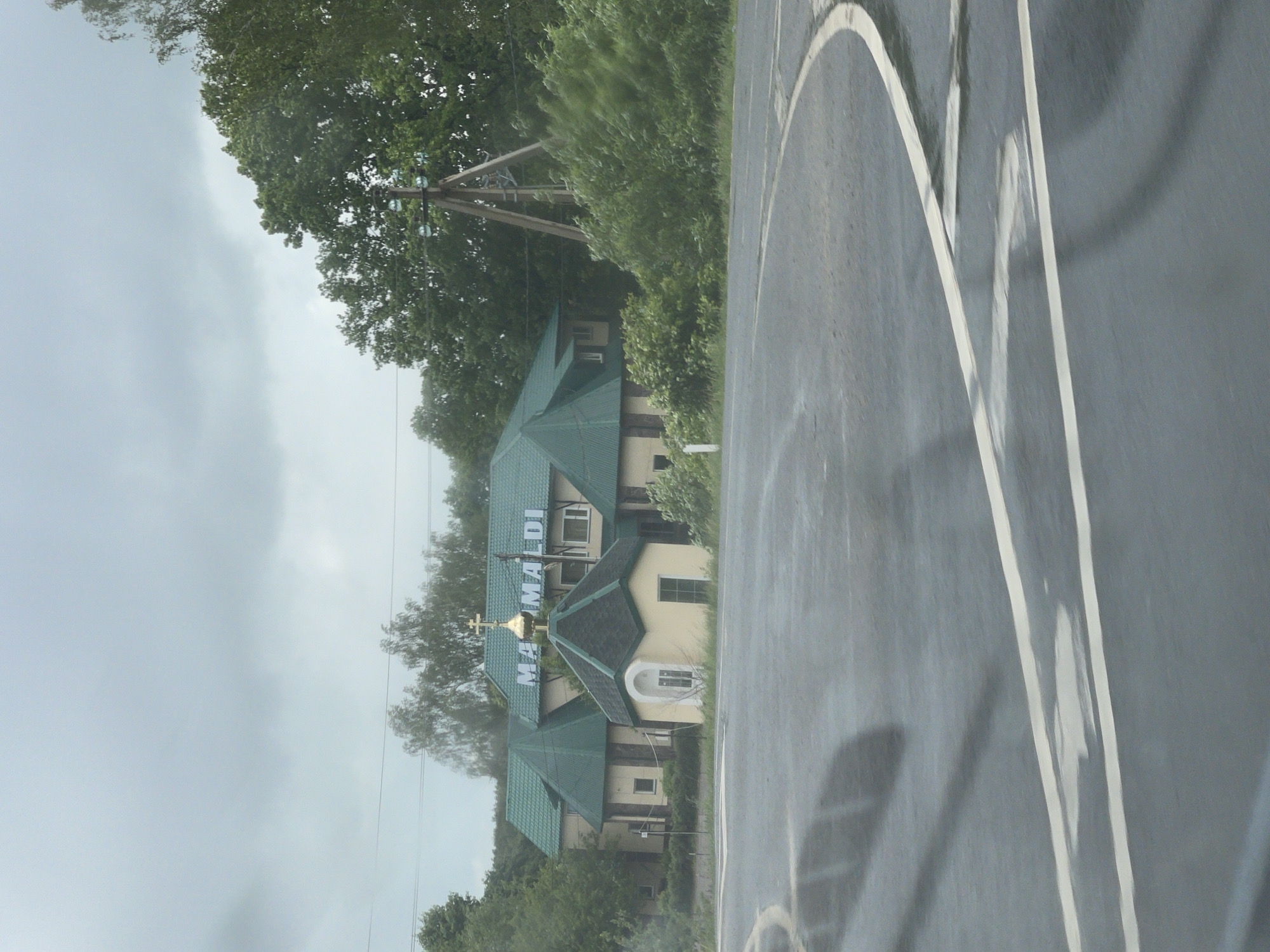 The image shows a view from inside a vehicle looking out onto a road with the "Mama Maldi" building visible. The building has a green roof, a gold cross, and an external sign that says "Mama Maldi" in blue lettering. The location is Кокино, Russia. The sky is overcast. Power lines and trees are also visible. White lines are painted on the road surface. The image is slightly rotated.