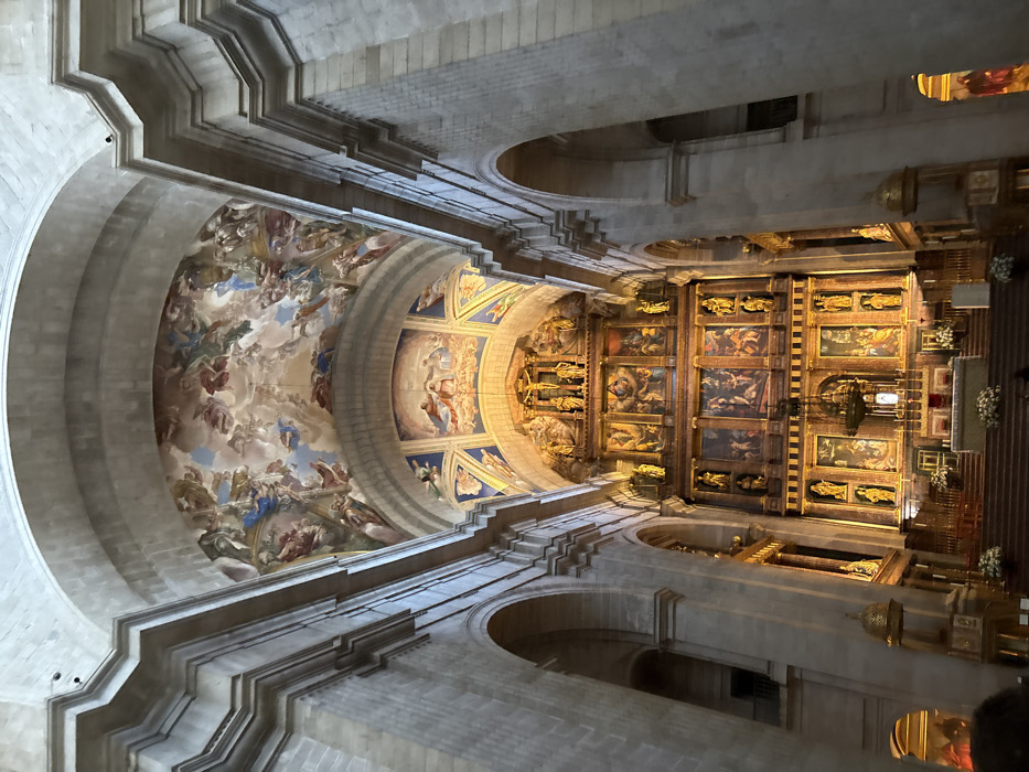The image depicts the interior of a grand religious edifice, specifically the Basilica within the Royal Monastery of San Lorenzo de El Escorial, located in San Lorenzo de El Escorial, Spain. The perspective is from the nave, looking directly towards the high altar and reredos.
The environment is characterized by massive, light-colored stone architecture, including soaring vaulted ceilings and large, rounded arches. The uppermost vaulted ceiling features an extensive fresco painting depicting numerous celestial figures, angels, and divine beings against a background of clouds and sky. Below this, a secondary, smaller vaulted section, directly above the reredos, is adorned with additional frescoes displaying religious figures and scenes, illuminated by a warm, artificial light source.
The focal point is the monumental, multi-tiered reredos (altarpiece) constructed from dark wood with intricate gilded ornamentation. The reredos is densely populated with numerous framed paintings depicting various religious narratives, interspersed with golden statues of saints or biblical figures housed in niches. At the apex of the reredos, a prominent crucifix with a sculpture of Christ is centered, flanked by more golden statues.
Below the reredos, a white altar table is visible, adorned with white floral arrangements and several candlesticks. A large, ornate chandelier hangs centrally above the altar. Multiple wide steps, covered in a dark material, lead up to the altar area, also featuring additional white floral arrangements. A low, dark barrier or railing separates the altar steps from the main floor of the nave.
The nave's side walls are lined with imposing stone pillars and arches, suggesting access to side aisles or chapels. A dark railing, possibly part of an upper gallery or balcony, is visible through an archway on the left. In the lower left foreground, a dark, indistinct silhouette of a person's head and shoulders is visible, indicating an observer of the scene. No specific actions or interactions are discernible beyond this implied observation.