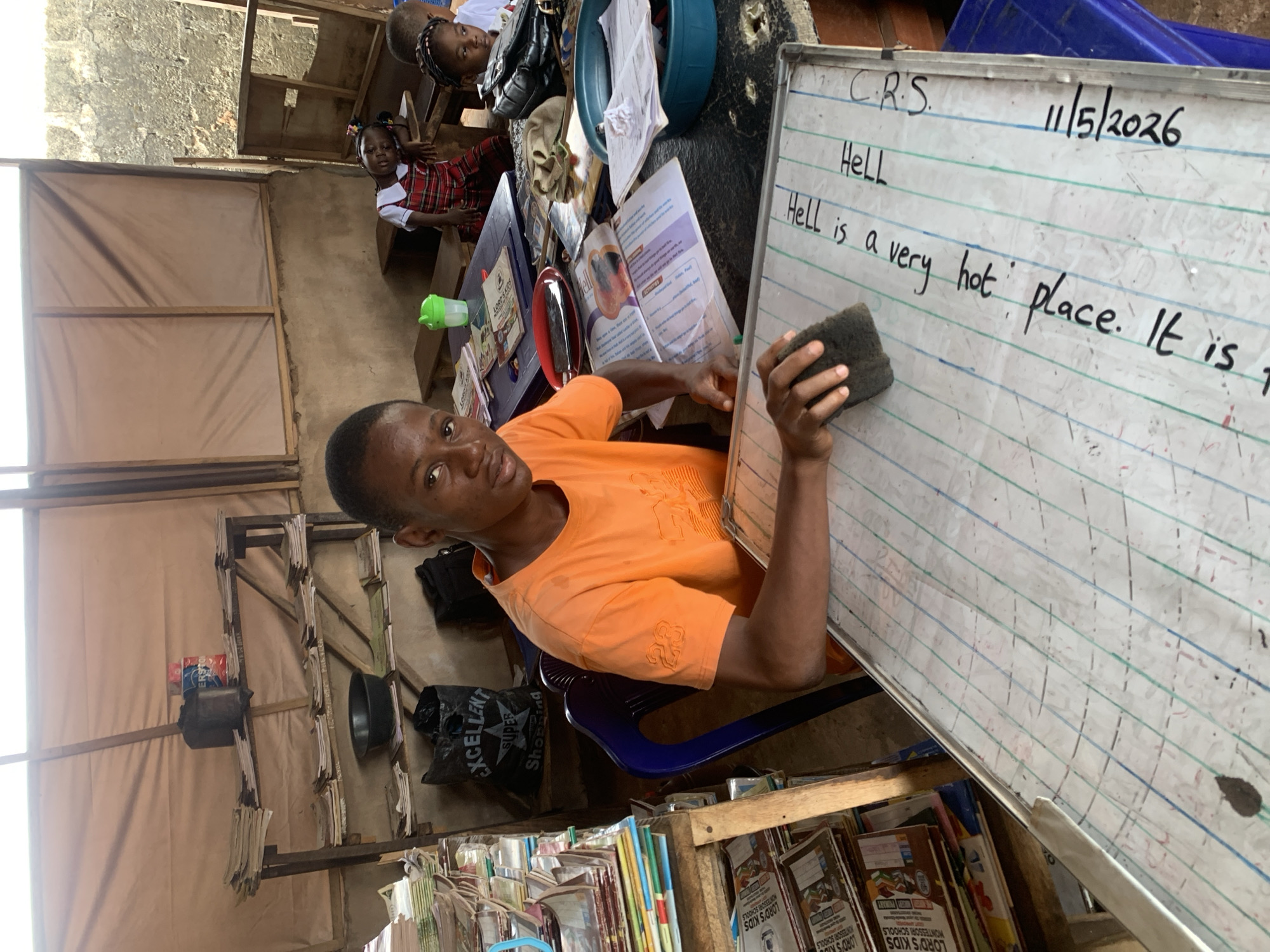 A young man wearing an orange t-shirt is erasing a whiteboard with the word "Hell" written on it, in what appears to be a classroom. The whiteboard includes the text: "C.R.S. 11/5/2026 Hell is a very hot place. It is".
Behind the man are two children sitting at a table, along with books and a washbasin. There is a rack filled with books and other items beneath the whiteboard. The "EXCELLENT SUPER Shopping" bag is nearby. The environment looks like a small classroom located in Ukwu-Abwa, Nigeria, filled with educational materials and supplies.