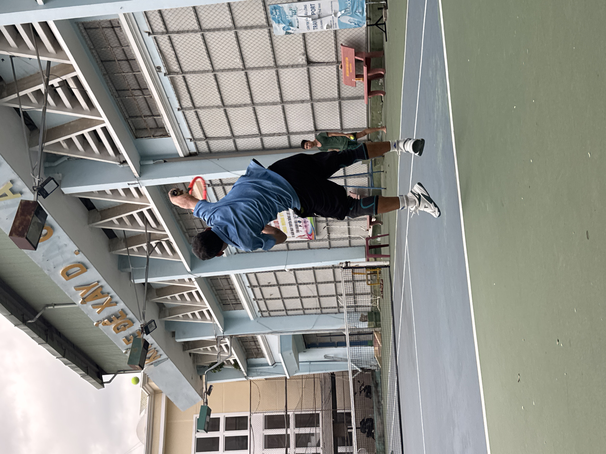 The image shows a tennis player in mid-air during a serve on an outdoor tennis court, likely at the Long Thọ Tennis Club in Nha Trang, Vietnam, given the building's architecture and signage. The player, wearing a blue shirt and black shorts with knee braces, is in the process of hitting a tennis ball. Another player in a green shirt stands in the background, holding a tennis racket. The court has a blue and green surface with white lines, and a net divides the two sides. In the background, a light blue building with prominent signage, possibly indicating a sports facility or school, is visible. There is also a red bench and a sign with Vietnamese text. The sky is overcast.
