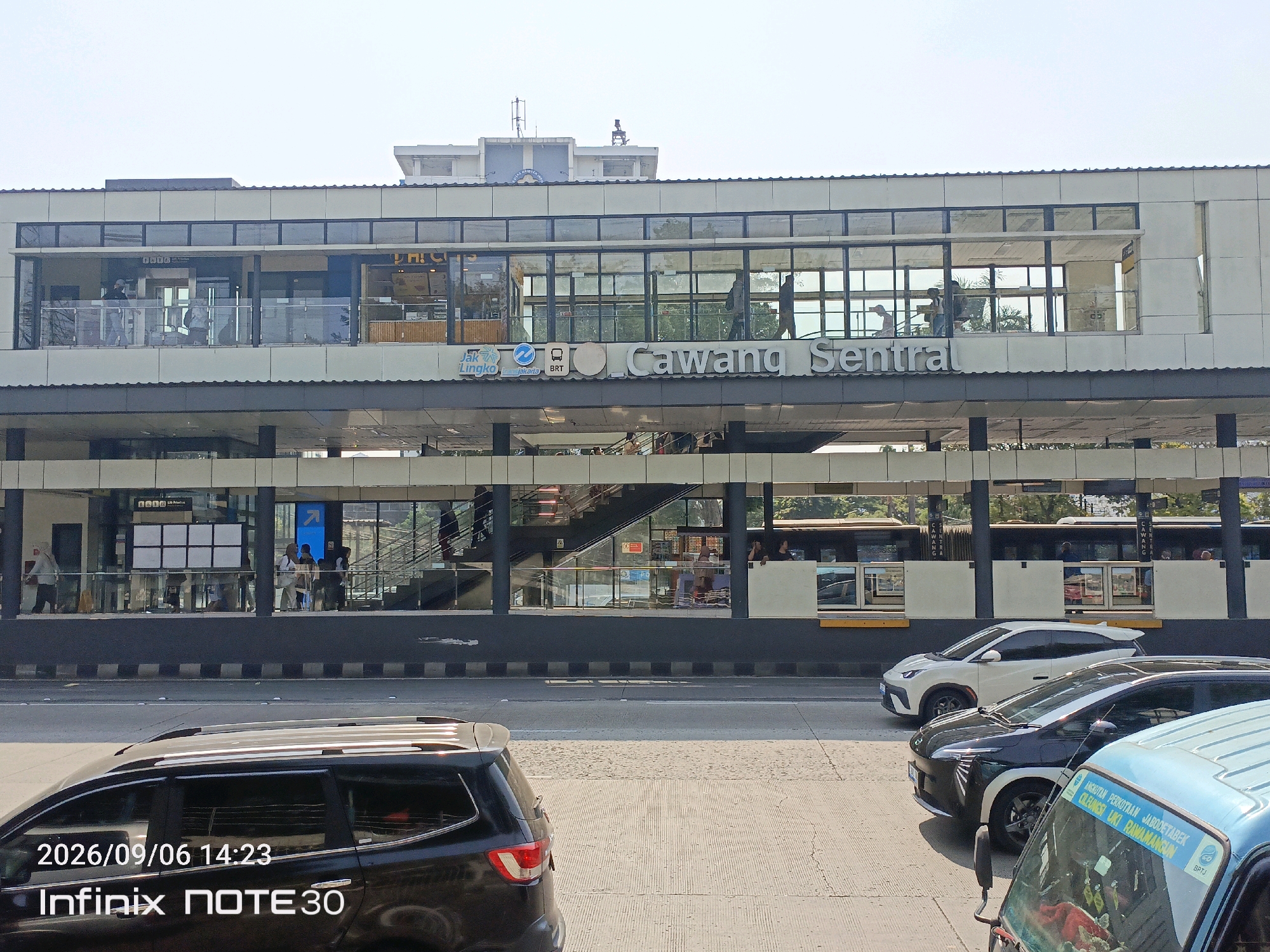 The image captures a street-level view of the Cawang Sentral Transjakarta bus stop in East Jakarta, Indonesia. The bus stop is a multi-level structure with a modern design featuring large glass windows and a prominent sign reading "Cawang Sentral." Several buses are visible behind the glass, indicating active service. On the street in the foreground, a black SUV is parked, and to its right, a light blue vehicle is partially visible. The sky is clear, suggesting daytime. The timestamp "2026/09/06 14:23" and "Infinix NOTE30" are overlaid on the bottom left of the image, likely originating from the camera's metadata.