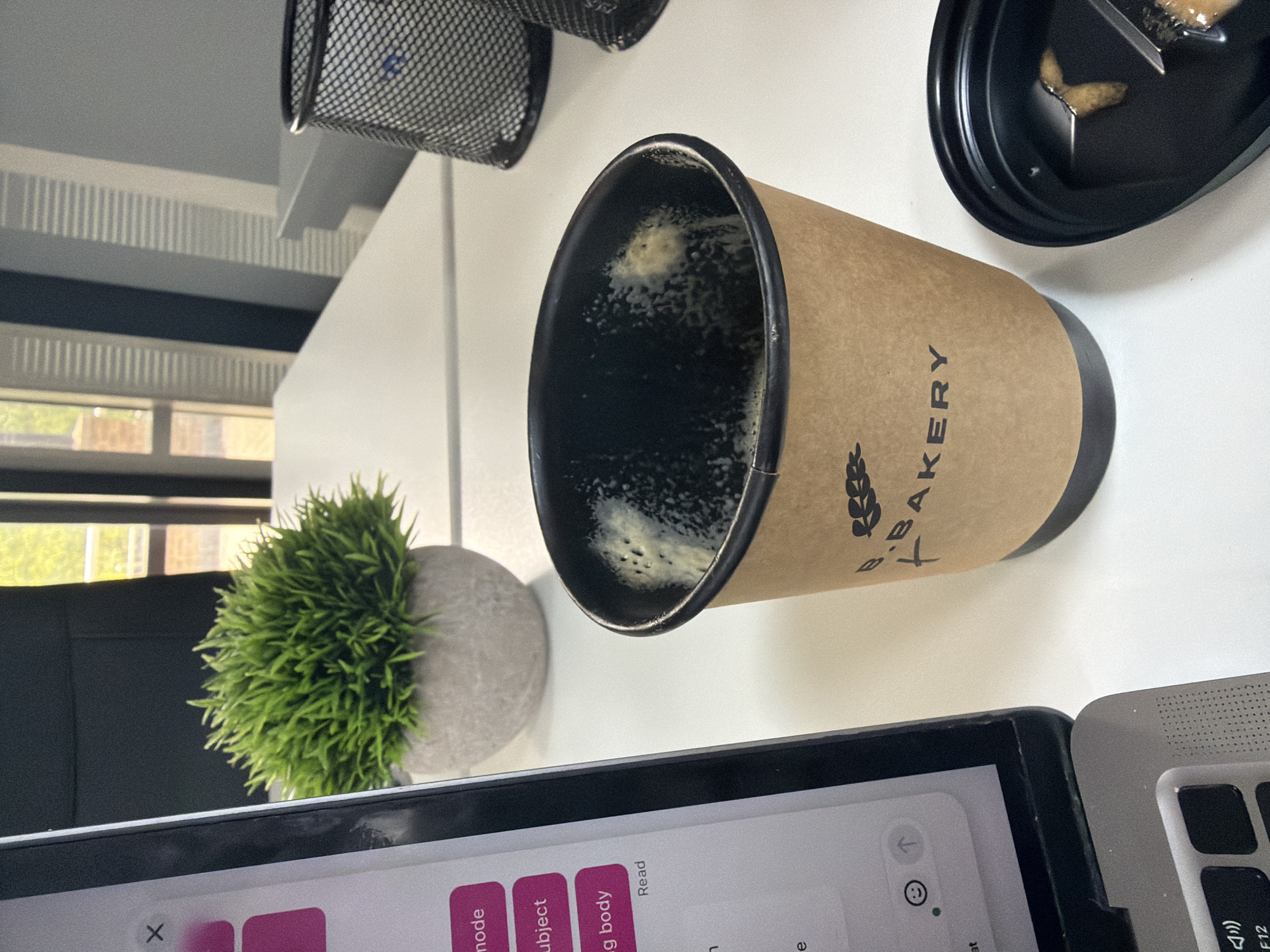 A high-angle view of a desk surface features a coffee cup from "T Bakery" with remnants of coffee inside. To the left, a small potted green plant sits near a black mesh container in the background. A laptop and a tablet are placed on the lower part of the desk, and a black container with food is in the top-right corner. The environment suggests an indoor workspace with natural light coming through a window. The location is presumed to be London, United Kingdom based on the Bakery name.