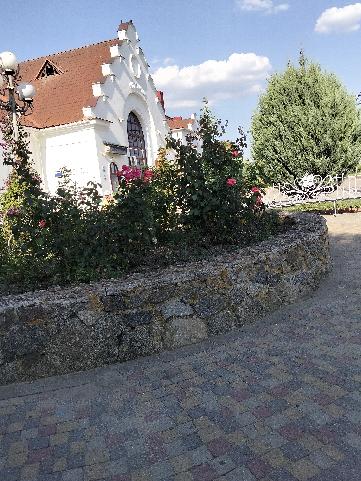 The image depicts the exterior of the Myrhorod train station in Myrhorod, Ukraine, on a sunny day. A white building with a red-tiled, gabled roof and decorative architectural elements, including a large arched window and a small triangular dormer, is visible. In front of the building, a curved stone planter bed overflows with lush green rose bushes in bloom, displaying vibrant pink and red flowers. A decorative white metal fence and a tall, dense evergreen tree stand behind the rose bushes. The foreground features a paved walkway with a colorful, geometric pattern of interlocking bricks in shades of grey, red, and pink. A traditional lamppost with multiple lights stands to the left of the building. The sky is blue with scattered white clouds.