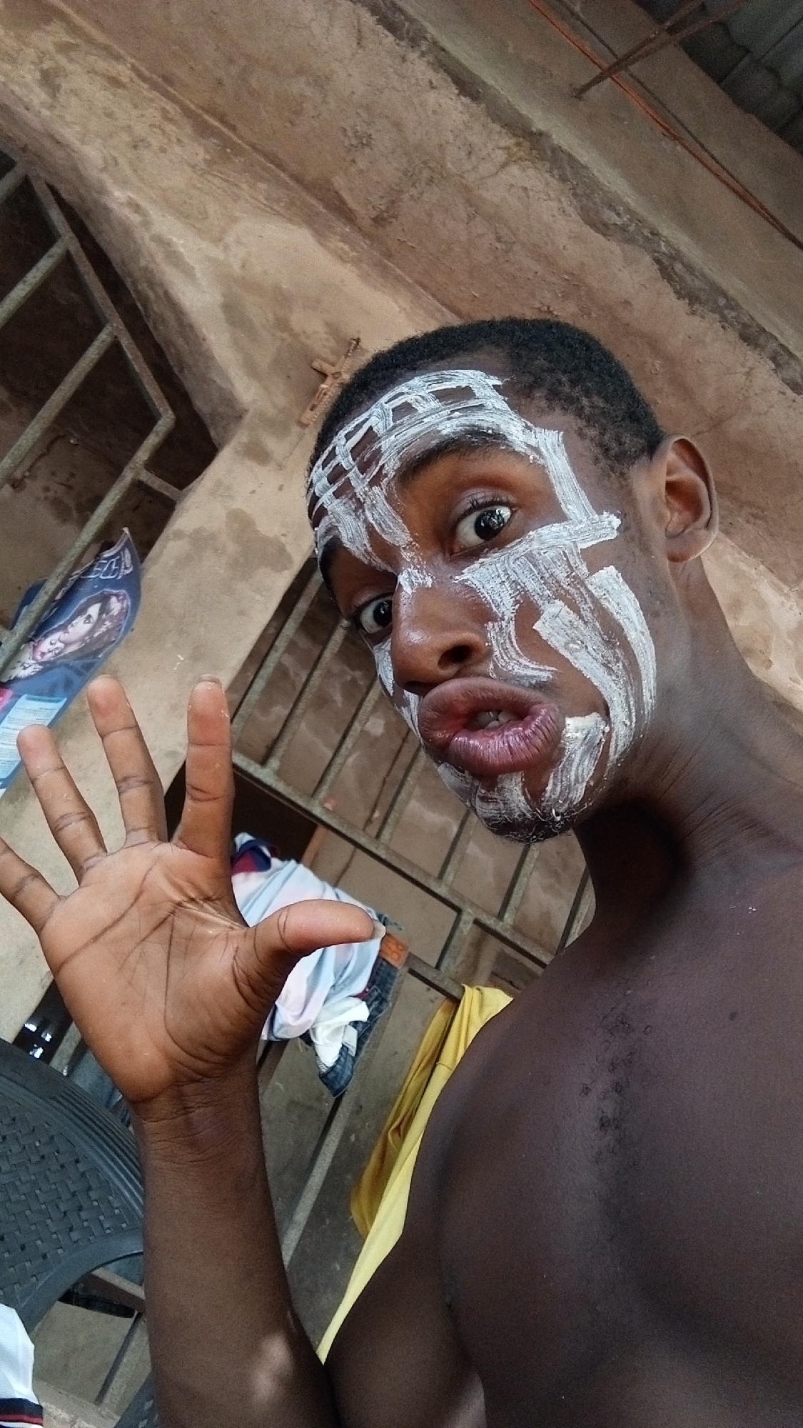 A young Black male is captured in a close-up, slightly angled shot. He has short, dark hair and is shirtless, revealing his bare chest and shoulders. White paint or markings are applied to his face and forehead in striking patterns, resembling stripes and geometric shapes. He is looking directly at the camera with wide, expressive eyes and puckered lips, conveying a playful or theatrical emotion. His left hand is raised in front of him, fingers spread open.
The background suggests an informal or rustic setting, possibly indoors or in a covered outdoor area. To the left, a dark, woven plastic chair is partially visible, along with some folded clothing (white, blue, and red patterns) on a surface. Behind the subject's right arm, a yellow garment is draped. The main background consists of uneven, concrete-like walls and a structure with horizontal and vertical metal bars, possibly a window grill. A patterned bag or fabric, featuring an image of a person (appears to be a woman with long dark hair), is visible behind his raised hand. A small, cross-shaped object is attached to the wall or a beam in the upper background. The lighting suggests it is daytime. No discernible text is visible in the image.