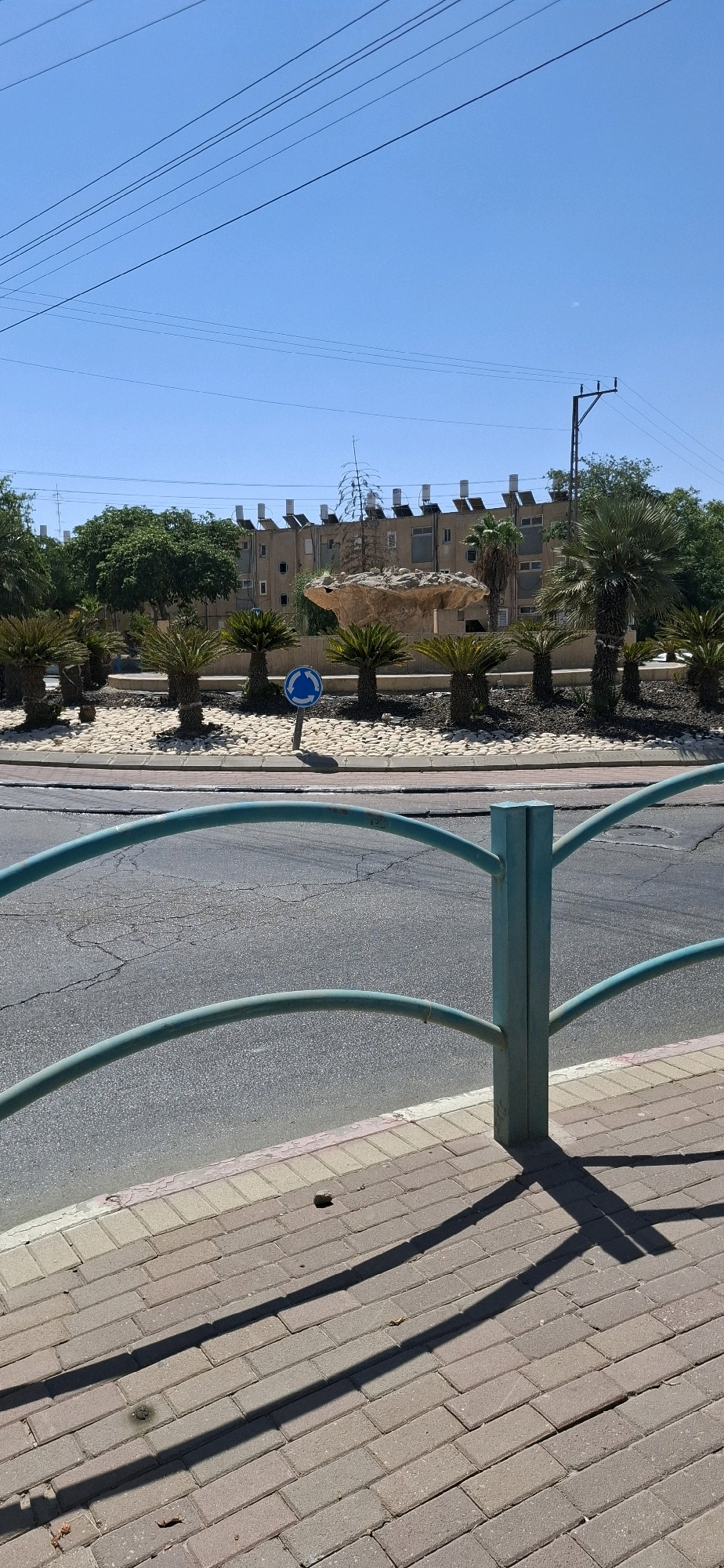 The image depicts a street scene in Dimona, Israel, on a sunny day. In the foreground, a turquoise metal railing with a vertical post separates the sidewalk from the road. The sidewalk is paved with light-colored interlocking bricks, and shadows cast by the railing fall across it. The road, with visible cracks, is in the middle ground. Beyond the road, a circular median is decorated with white pebbles, several small palm-like plants, and a blue roundabout sign. A large, rock-like sculpture is visible in the center of the median. In the background, a row of buildings with multiple windows and solar water heaters on their roofs lines the street. Trees and a utility pole are also present. The sky is clear and blue.
