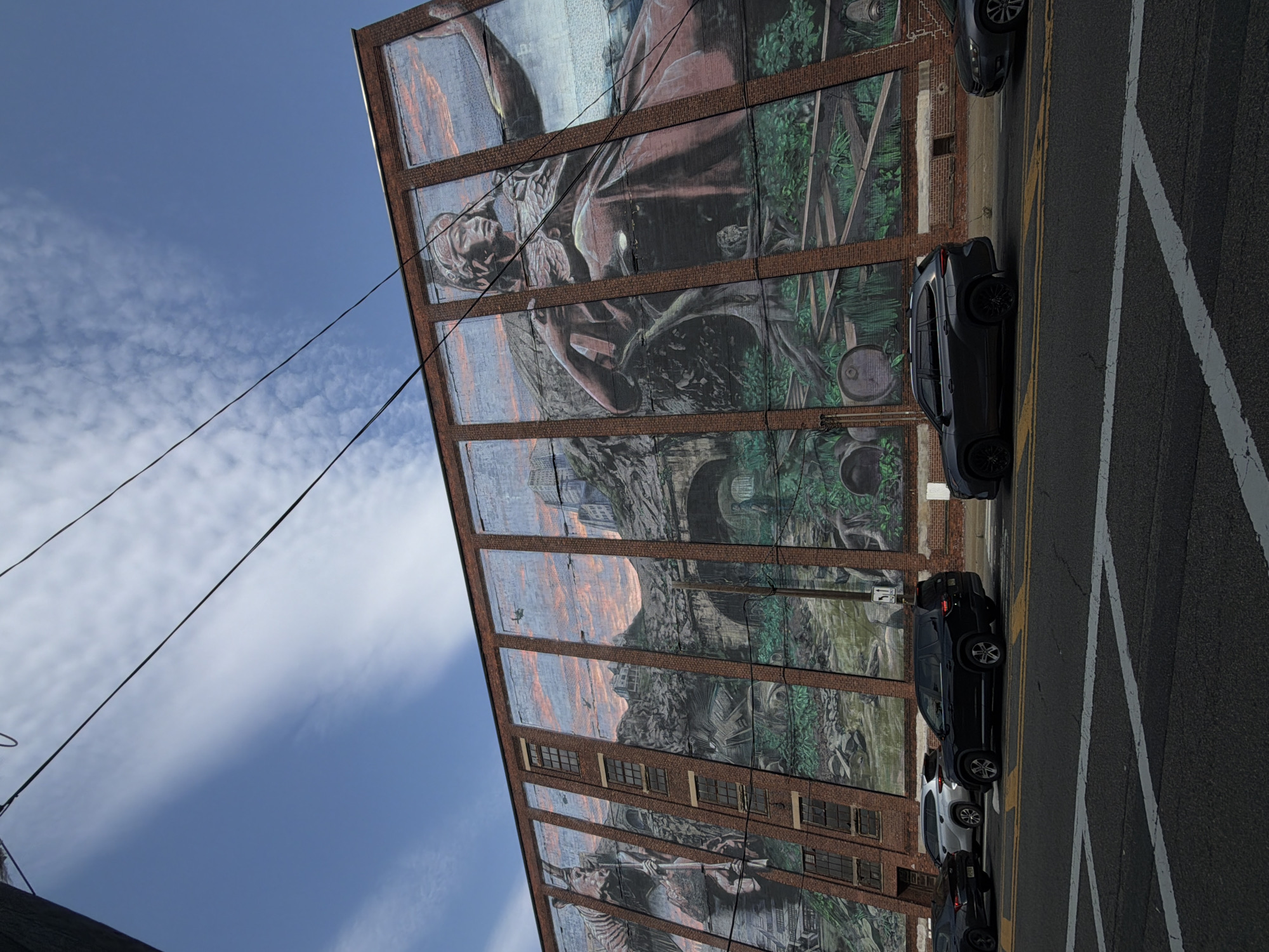 The image shows a large mural painted on the side of a brick building in Jersey City, United States of America. The mural depicts a scene with mountainous terrain, a large figure, and possibly industrial elements. The sky above is blue with scattered clouds, and power lines are visible crisscrossing the frame. In the foreground, several cars are parked or driving on a street that runs parallel to the building. The mural is divided into several vertical panels, each featuring a section of the overall artwork. The scale of the mural suggests it is a prominent feature in the urban landscape.