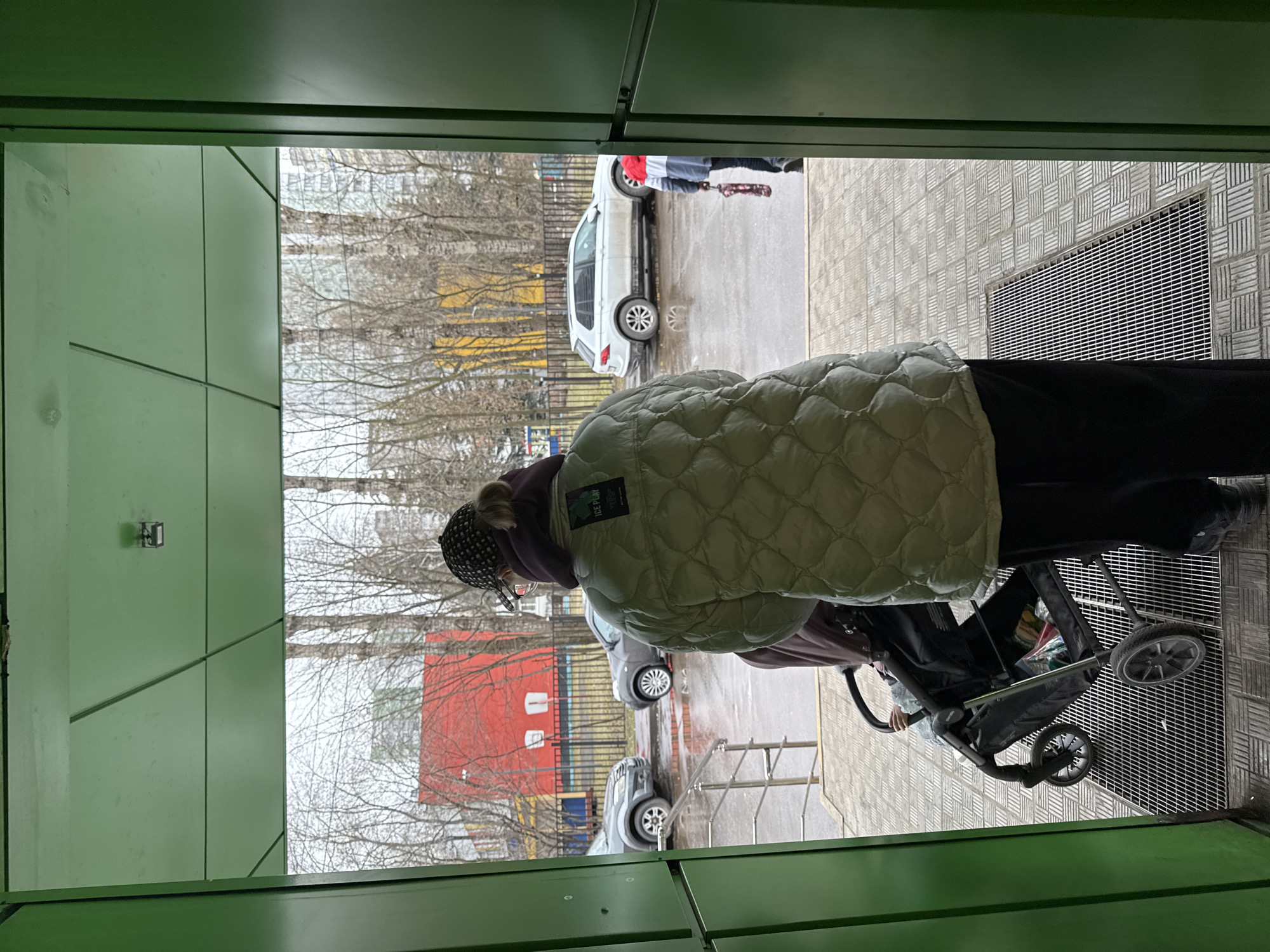 A person wearing a puffy jacket and pushing a stroller is entering a green doorway. Outside the doorway, a paved area is visible with several cars parked, as well as buildings painted yellow and red. The photograph was likely taken in the city of Moscow, Russia.