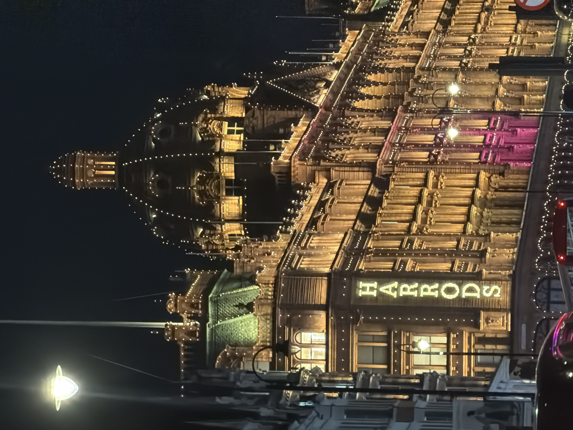 The image depicts the Harrods department store in London, United Kingdom, at night. The building is illuminated with numerous strings of lights, highlighting its architectural details and domes. The store's name, "HARRODS," is prominently displayed in illuminated letters on its facade. The environment is dark, suggesting it's nighttime, and streetlights are visible in the foreground.